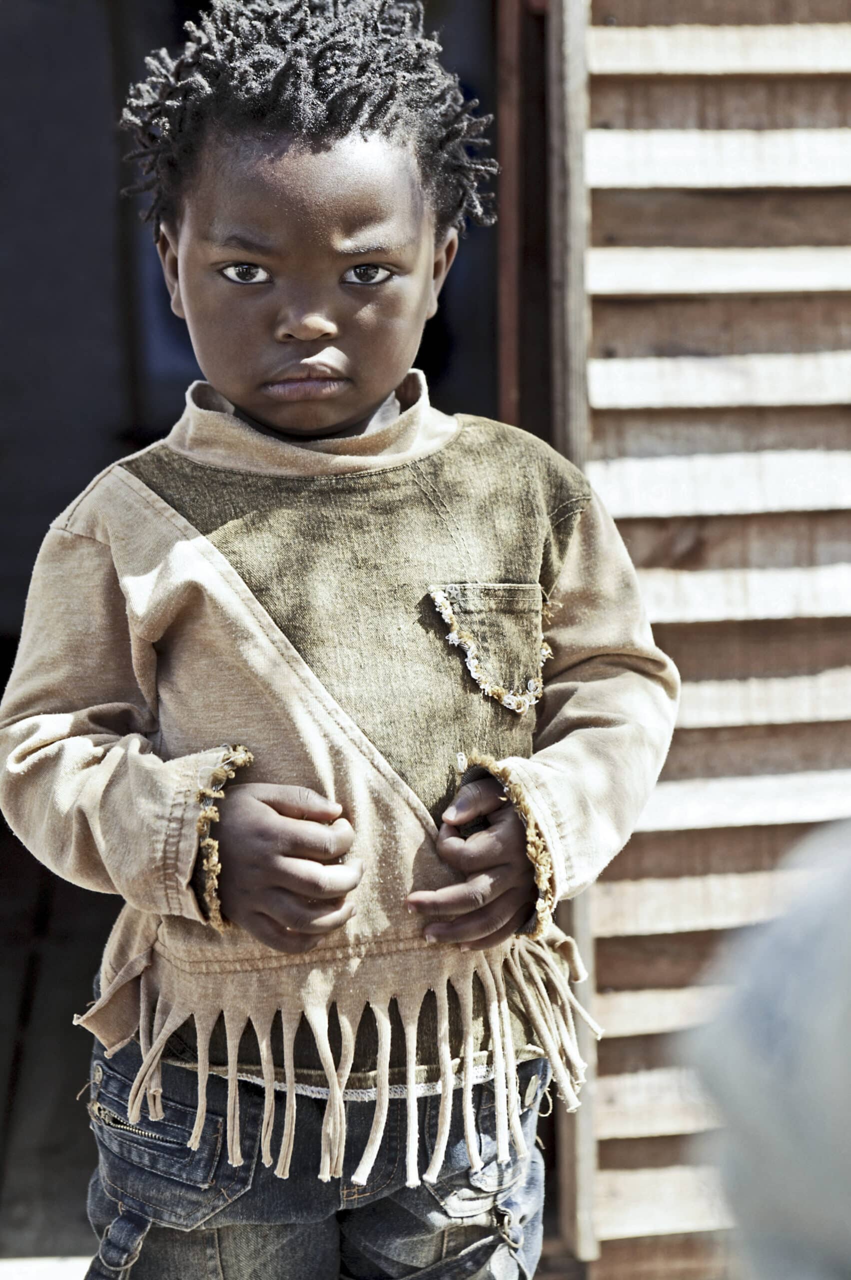Ein kleines Kind mit ernster Miene steht vor einem hölzernen Hintergrund. Das Kind trägt einen zerfetzten beigen Pullover und blaue Jeans und umklammert den Stoff des Pullovers. © Fotografie Tomas Rodriguez