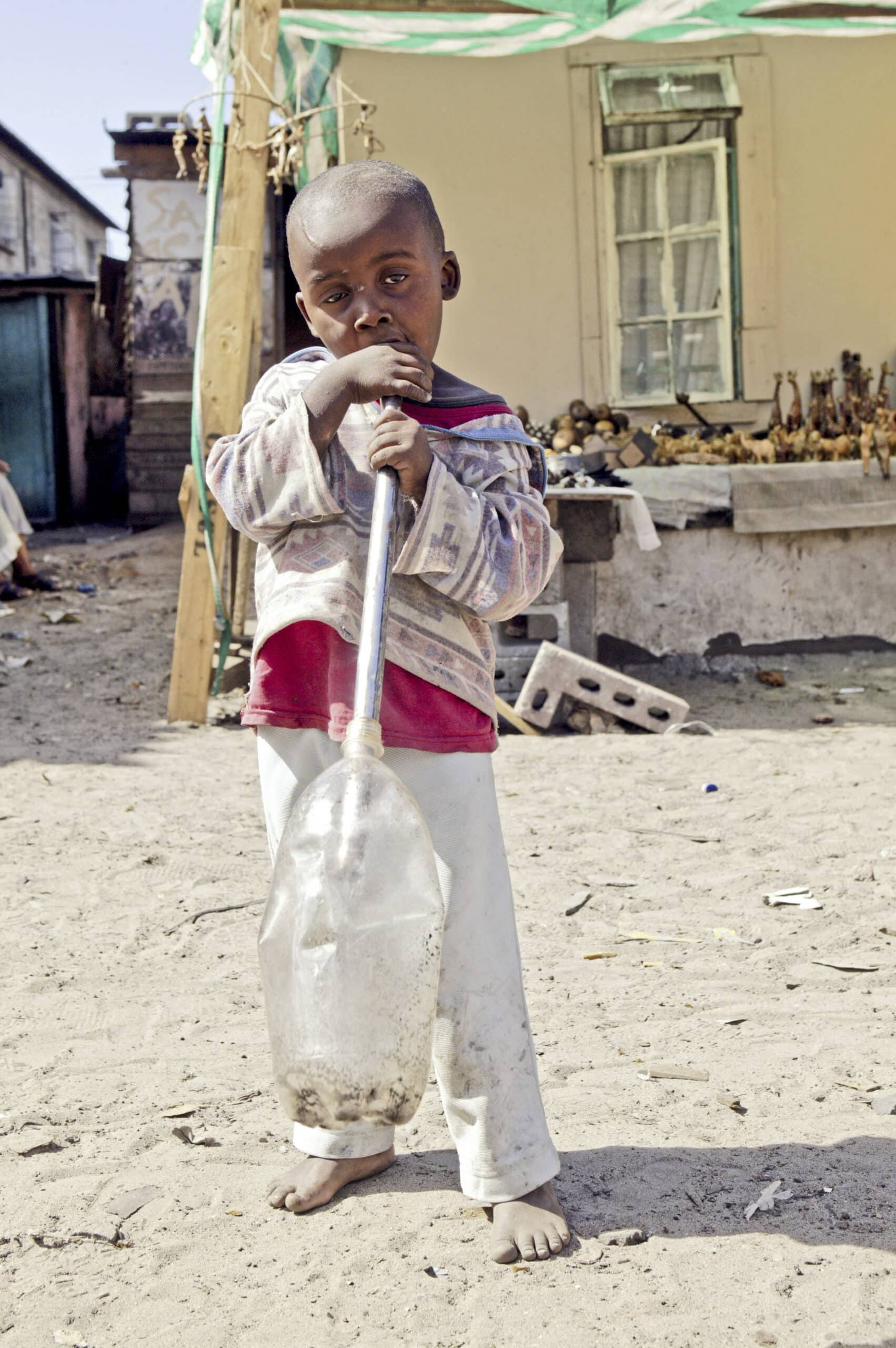 Ein kleiner Junge steht barfuß auf einem Sandboden und hält eine große Plastikflasche mit einem hindurchgesteckten Stock in der Hand. Im Hintergrund sind ein bescheidenes Haus und verschiedene kleine Gegenstände auf einem Tisch angeordnet. © Fotografie Tomas Rodriguez