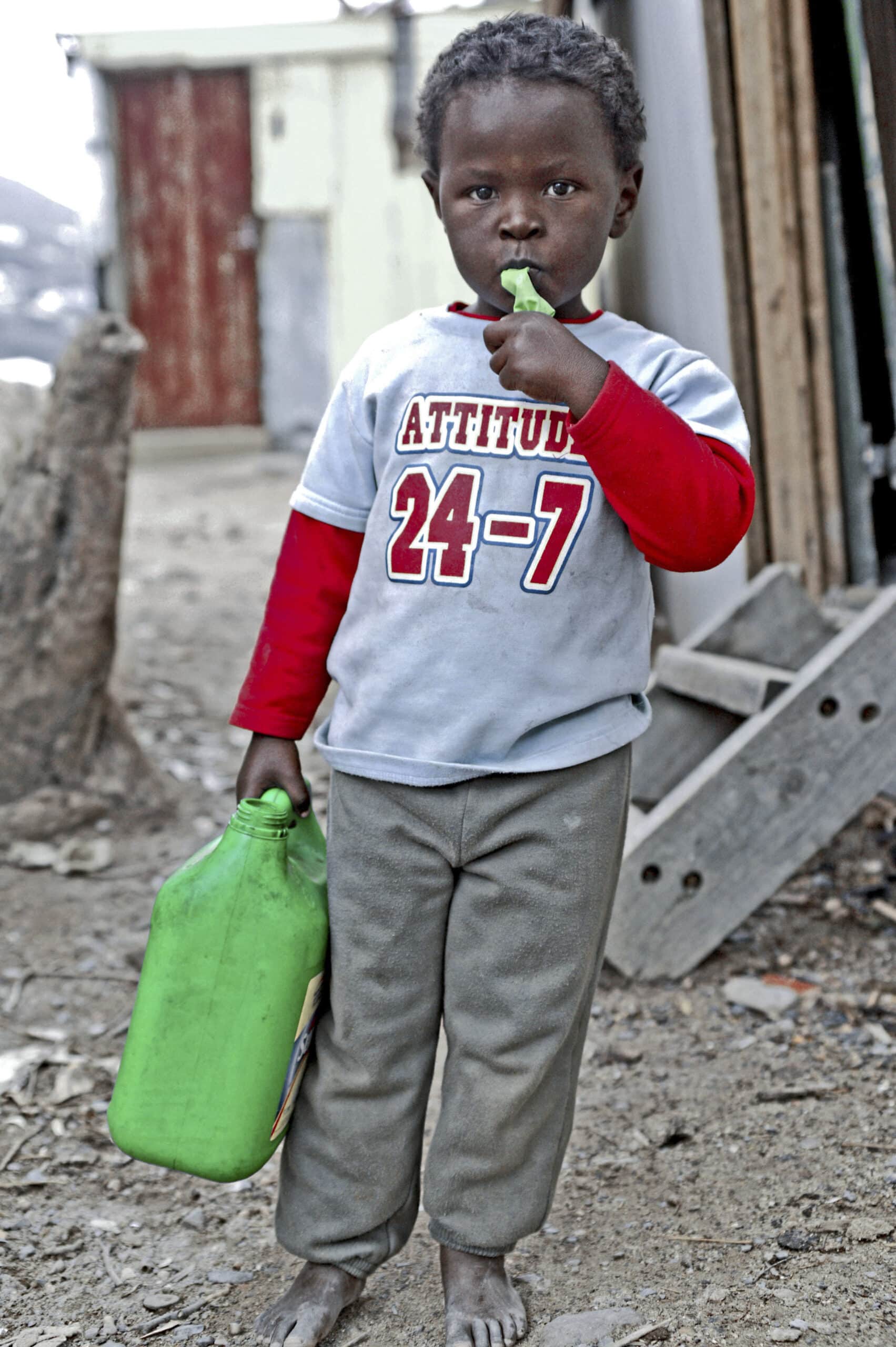 Ein kleines Kind steht im Freien, hält einen grünen Krug in der Hand und ein Blatt im Mund und trägt ein rot-weißes T-Shirt mit der Aufschrift „Attitude 24-7“ und eine graue Hose. © Fotografie Tomas Rodriguez