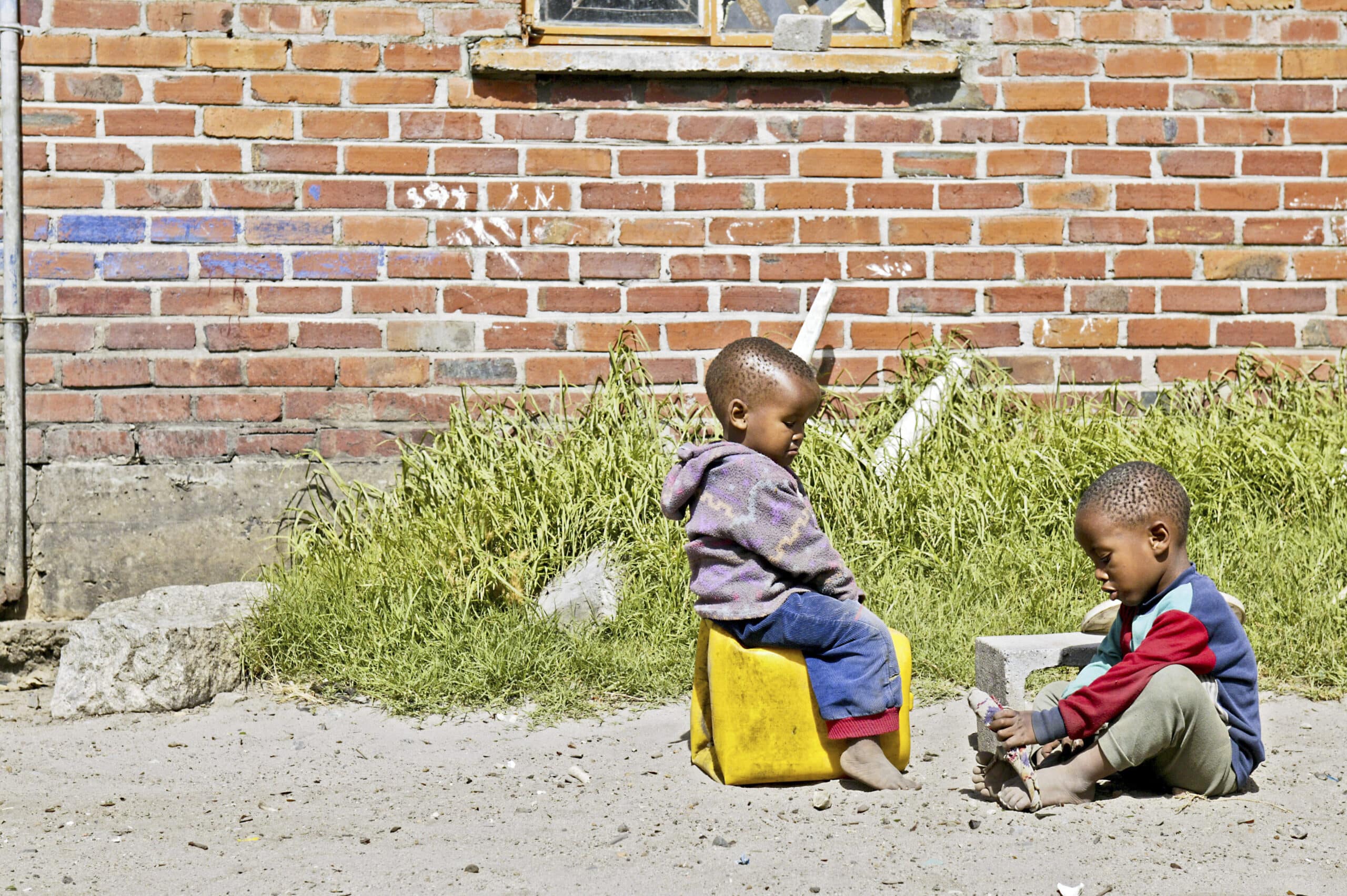 Zwei kleine Kinder spielen draußen neben einer Backsteinmauer mit Graffiti. Das eine sitzt auf einem gelben Eimer, das andere auf dem Boden und ist in einen spielerischen Austausch verwickelt. © Fotografie Tomas Rodriguez