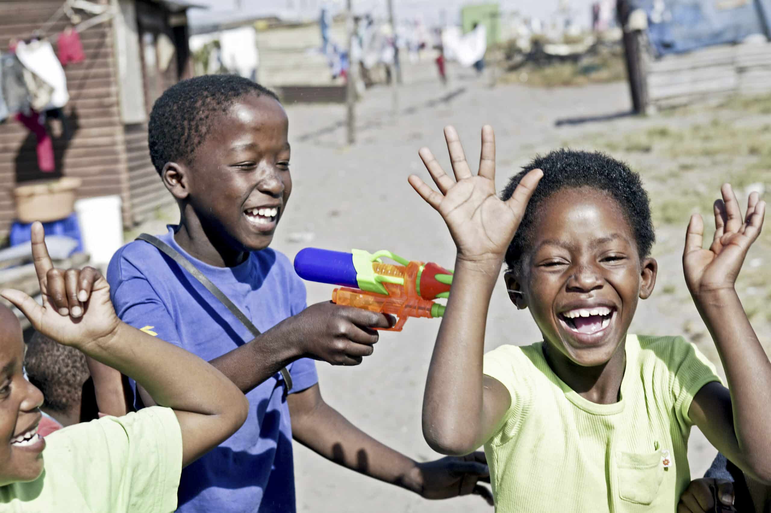 Drei Kinder spielen an einem sonnigen Tag in einer rustikalen Umgebung im Freien fröhlich mit einer Wasserpistole, und in ihren Gesichtern ist Lachen und Aufregung deutlich zu erkennen. © Fotografie Tomas Rodriguez