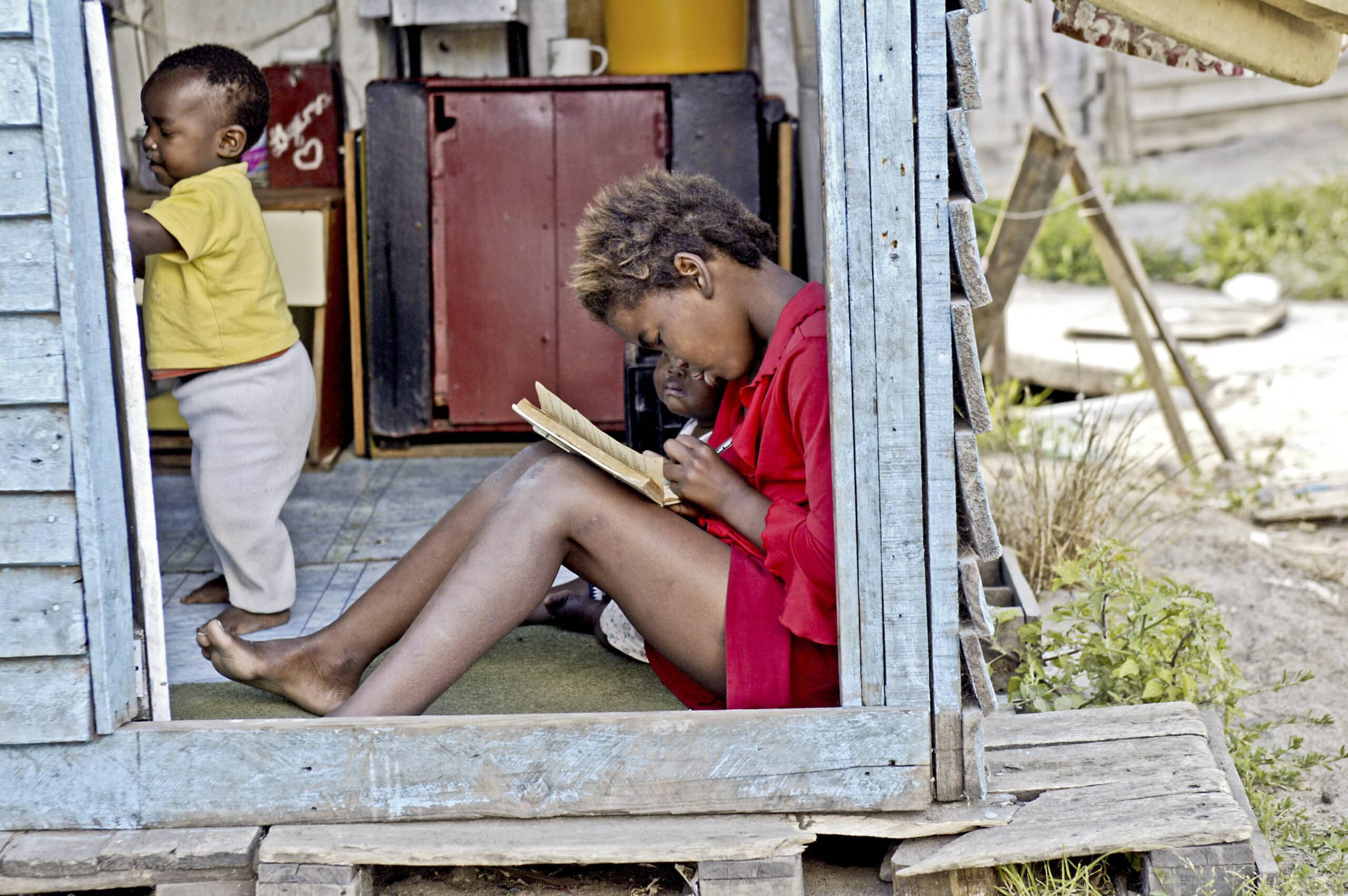 Ein junges Mädchen in einer roten Jacke sitzt im Eingang eines provisorischen Holzhauses und liest ein Buch. Im Hintergrund spielt ein Kleinkind neben einer offenen Tür. © Fotografie Tomas Rodriguez