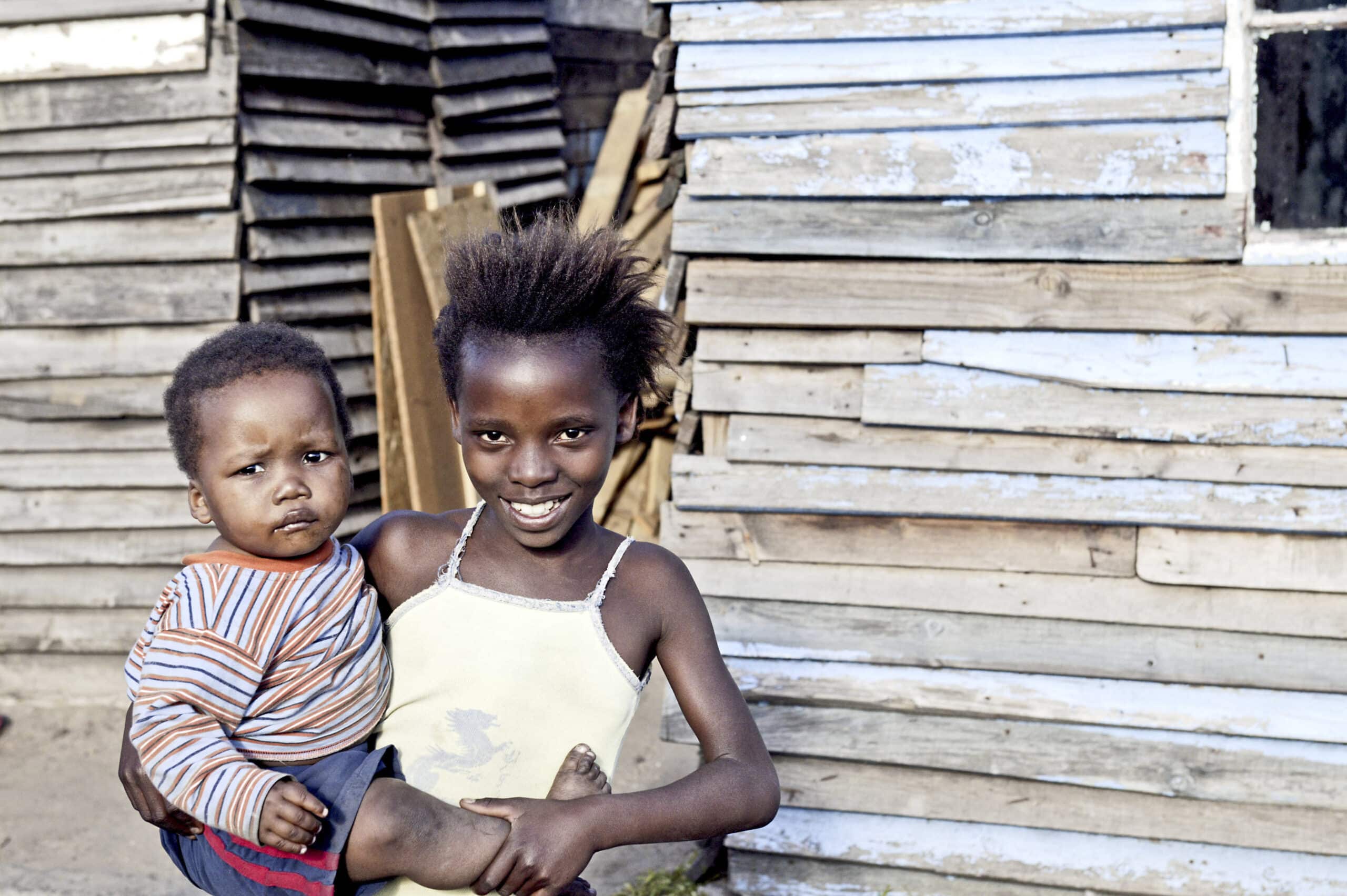 Ein junges Mädchen mit einem strahlenden Lächeln hält ein Kleinkind vor einer Holzhütte. Das Mädchen trägt ein gelbes Oberteil und das Kleinkind ein gestreiftes Hemd. Beide wirken eng und liebevoll. © Fotografie Tomas Rodriguez