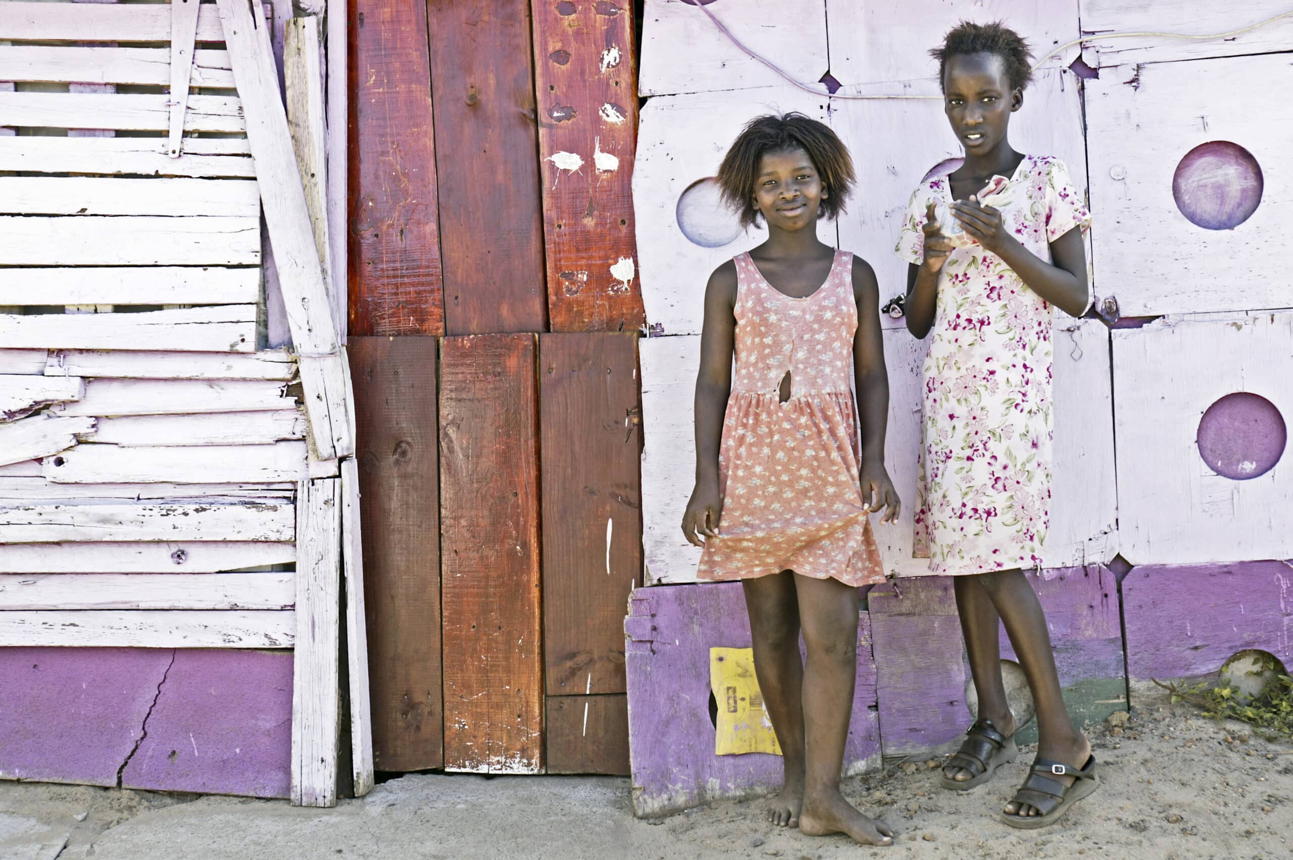 Zwei junge Mädchen stehen vor einem rustikalen Holzhaus mit lila und weißen Paneelen. Eines hält ein Buch, das andere eine Plastikflasche, beide barfuß auf sandigem Boden. © Fotografie Tomas Rodriguez
