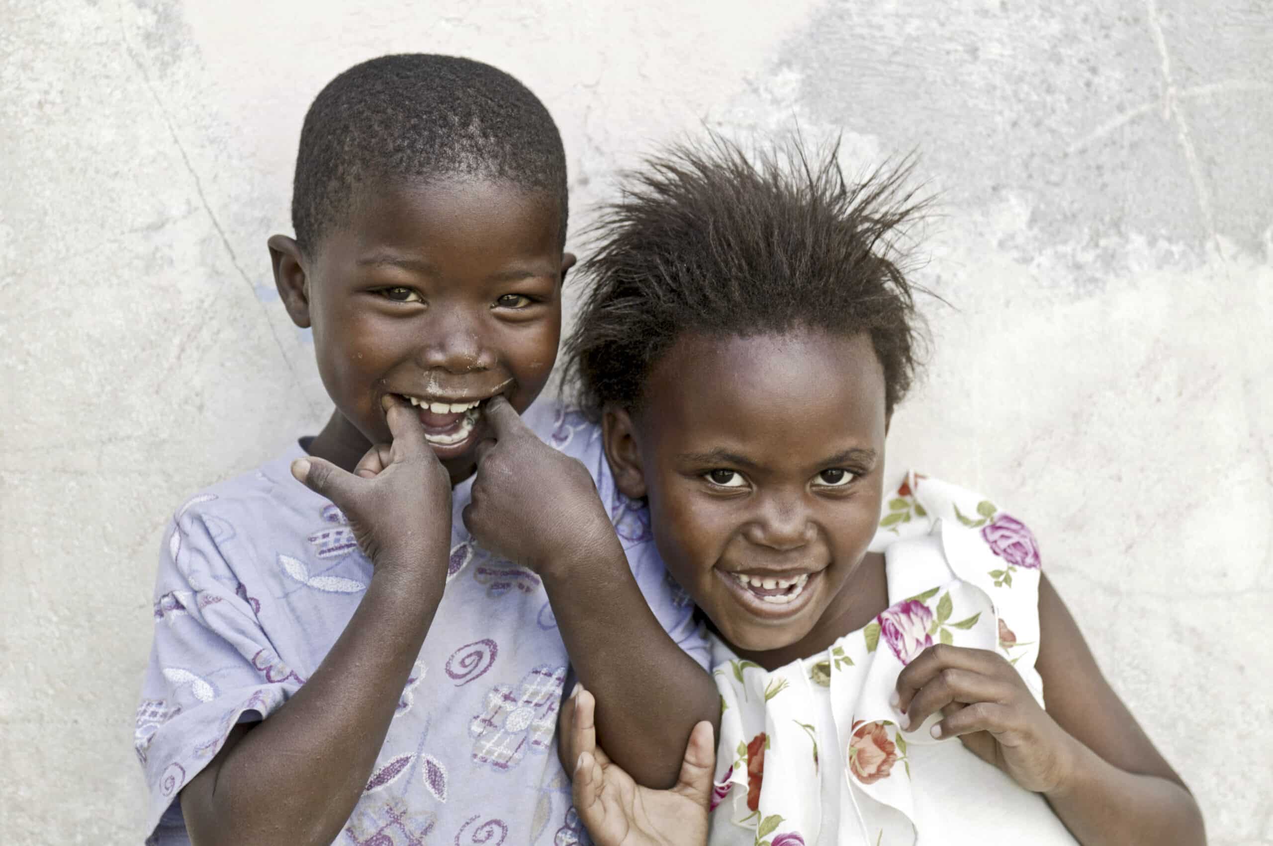 Zwei fröhliche Kinder lächeln und spielen, eines in einem helllila Hemd und das andere in einem weißen geblümten Kleid, vor einer strukturierten beigen Wand. © Fotografie Tomas Rodriguez