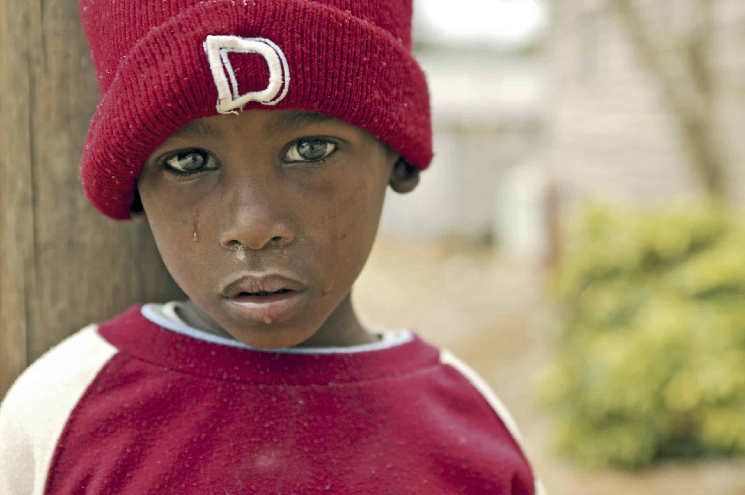 Ein kleiner Junge mit ernstem Blick und einer roten Mütze mit dem Buchstaben „D“ steht draußen und blickt direkt in die Kamera. Sein Gesicht zeigt Tränenspuren, die seinen ergreifenden Ausdruck noch verstärken. © Fotografie Tomas Rodriguez
