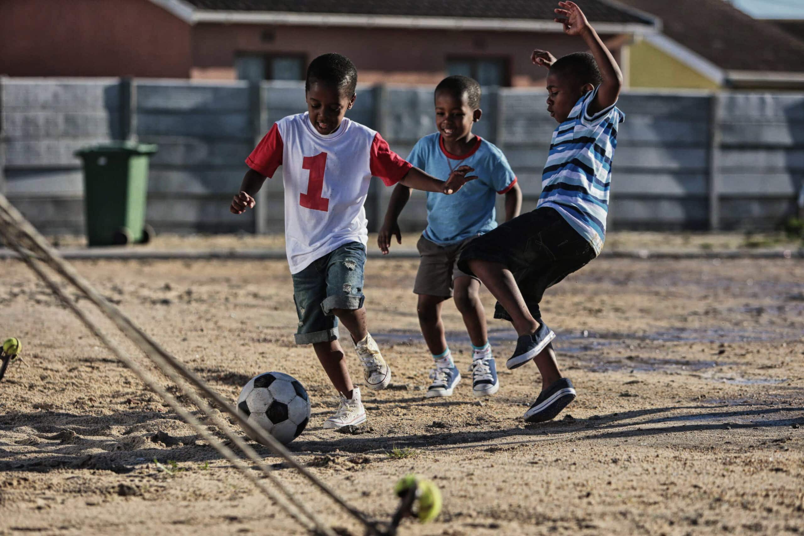Drei Jungen spielen Fußball auf einem Feld aus Erde. Ein Junge in einem weiß-roten Trikot mit der Nummer 1 kickt den Ball, während die anderen versuchen, ihn abzufangen. Sie scheinen das Spiel zu genießen. Im Hintergrund sind Häuser und eine grüne Mülltonne zu sehen. © Fotografie Tomas Rodriguez