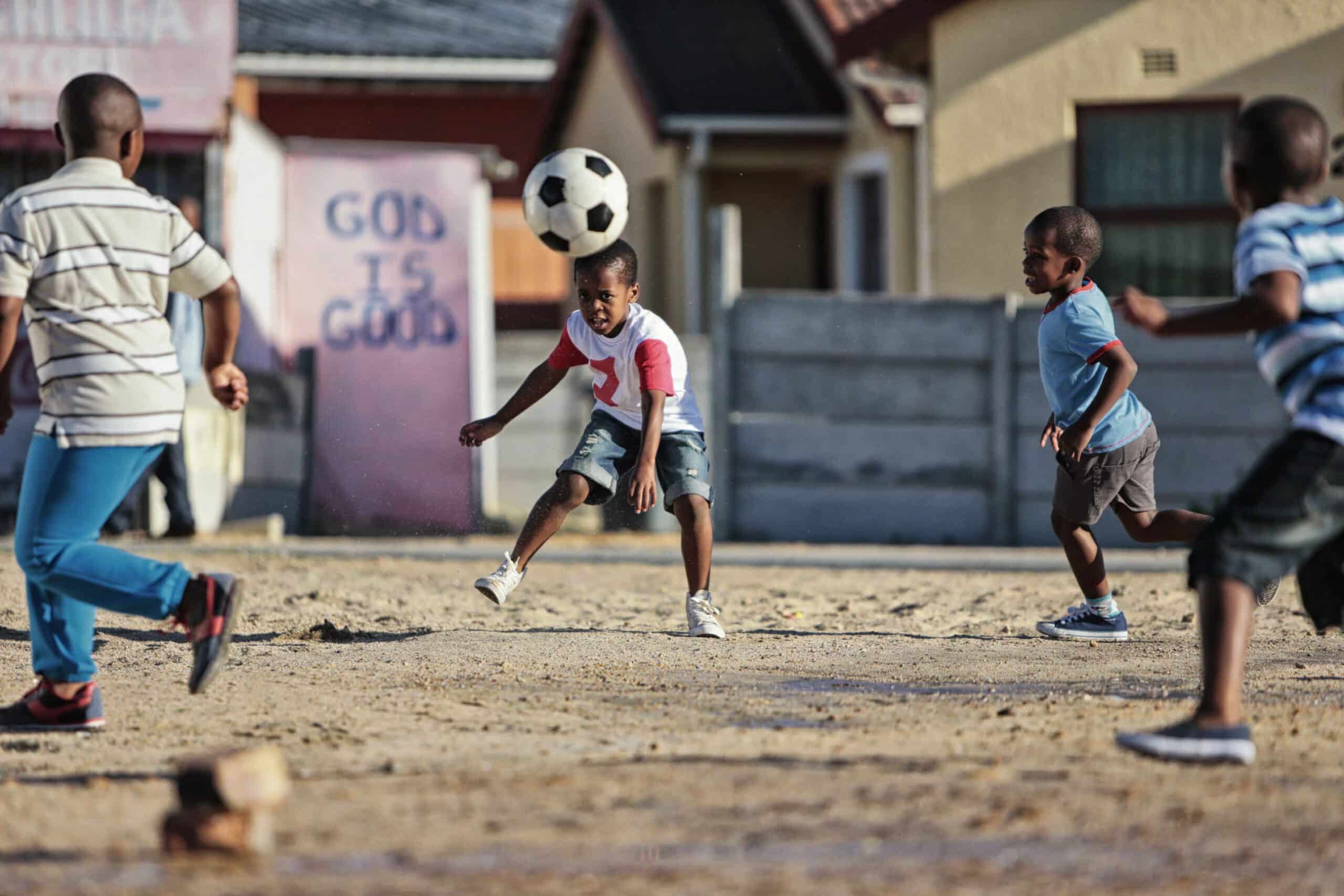 Vier junge Jungen spielen Fußball auf einem Feld in einem Wohngebiet. Ein Junge ist gerade dabei, einen Fußball zu köpfen, während die anderen Jungen rennen und zuschauen. Auf einer Wand im Hintergrund steht die Botschaft „GOTT IST GUT“. Im Hintergrund sind Häuser und Zäune zu sehen. © Fotografie Tomas Rodriguez