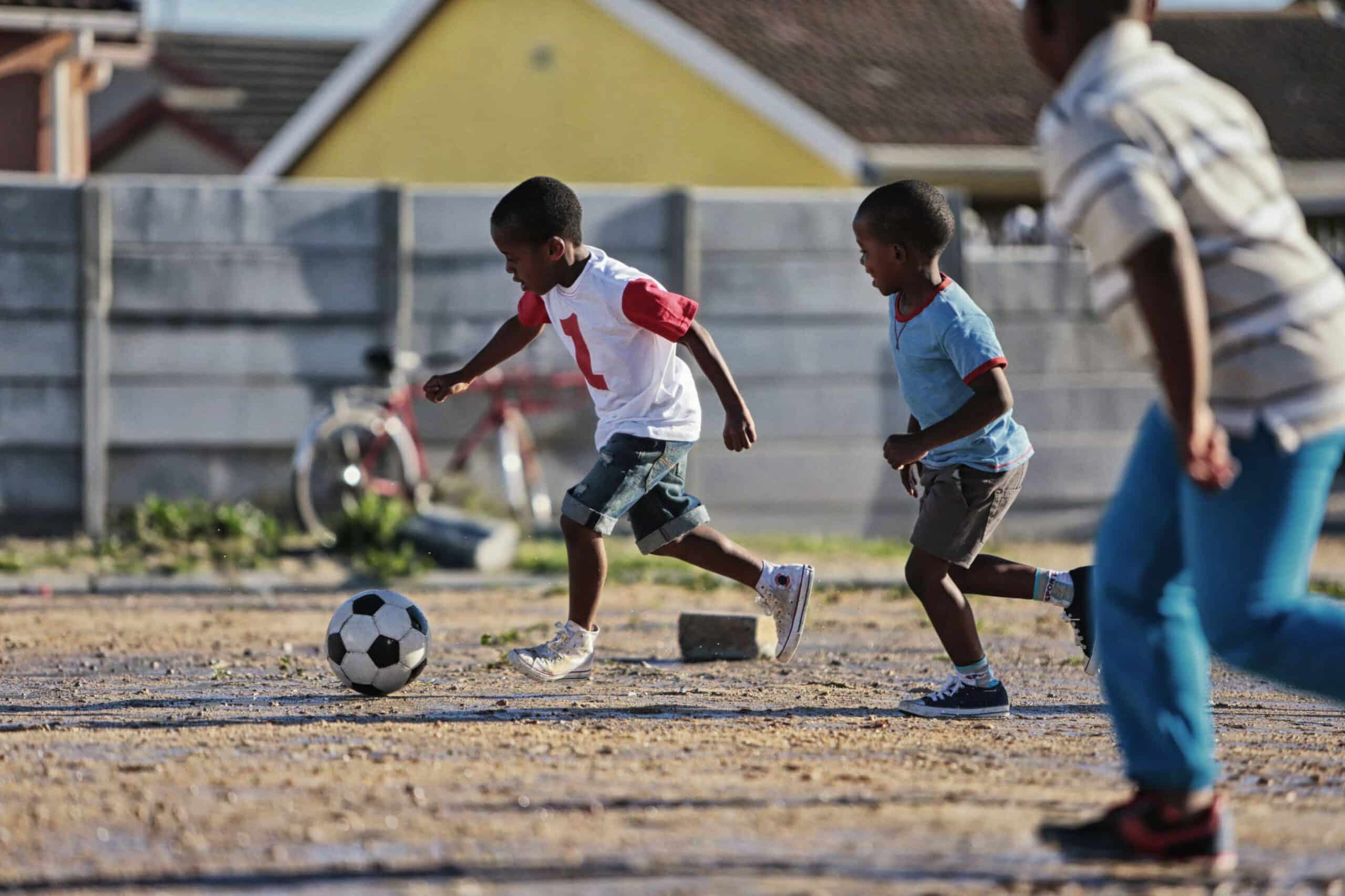 Drei Jungen spielen Fußball auf einem Feld aus Erde. Ein Junge, der ein weißes Trikot mit roten Ärmeln und der Nummer 1 trägt, jagt dem Ball hinterher, während die anderen beiden Jungen ihm dicht auf den Fersen sind. Im Hintergrund lehnt ein Fahrrad an einer Mauer, hinter der sich Häuser befinden. © Fotografie Tomas Rodriguez