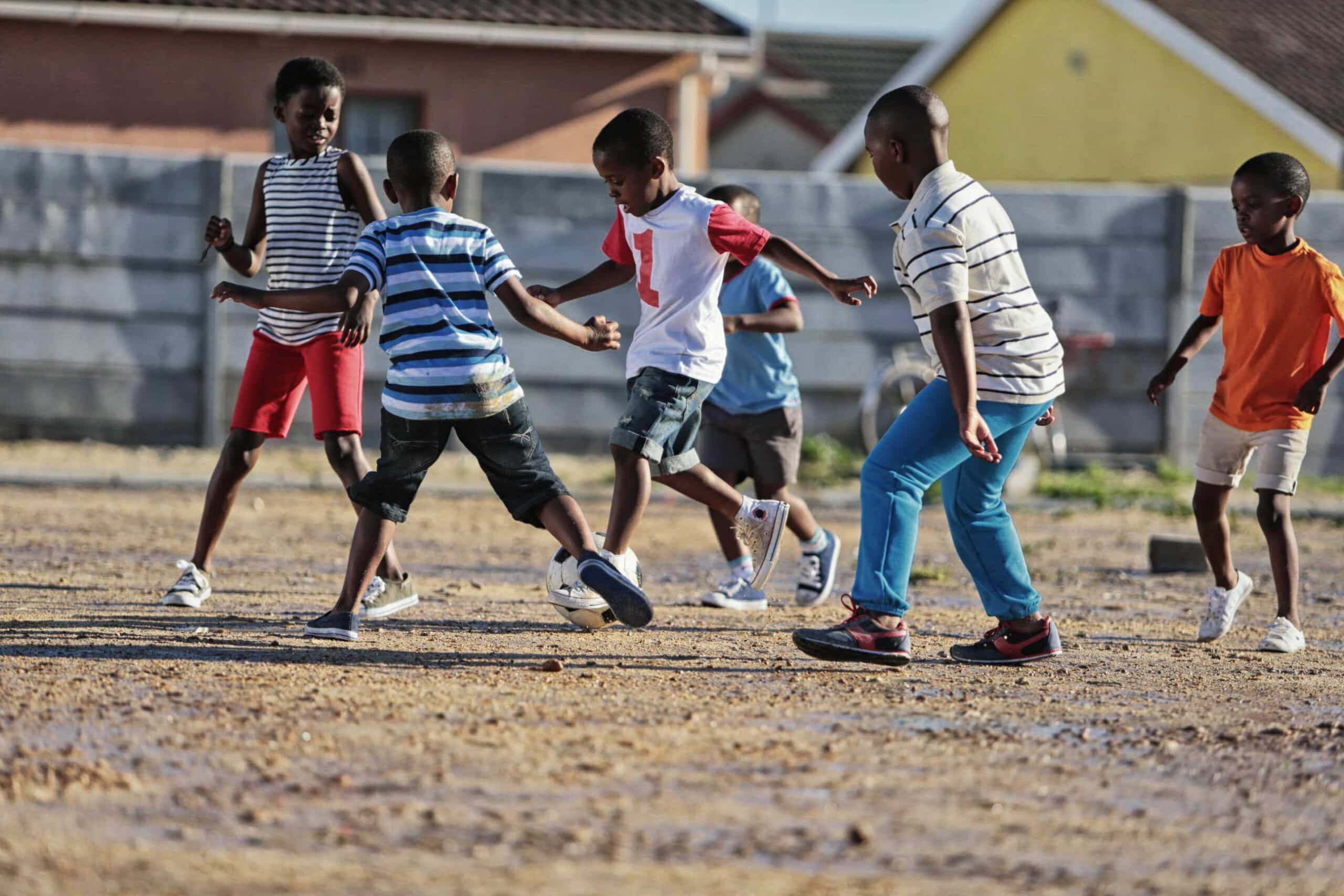 Eine Gruppe von Jungen spielt Fußball auf einem Feld aus Erde. Sie tragen Freizeitkleidung und jagen aktiv dem Ball hinterher. Im Hintergrund sind Häuser und ein Zaun zu sehen. Die Kinder scheinen das Spiel unter einem klaren Himmel zu genießen. © Fotografie Tomas Rodriguez
