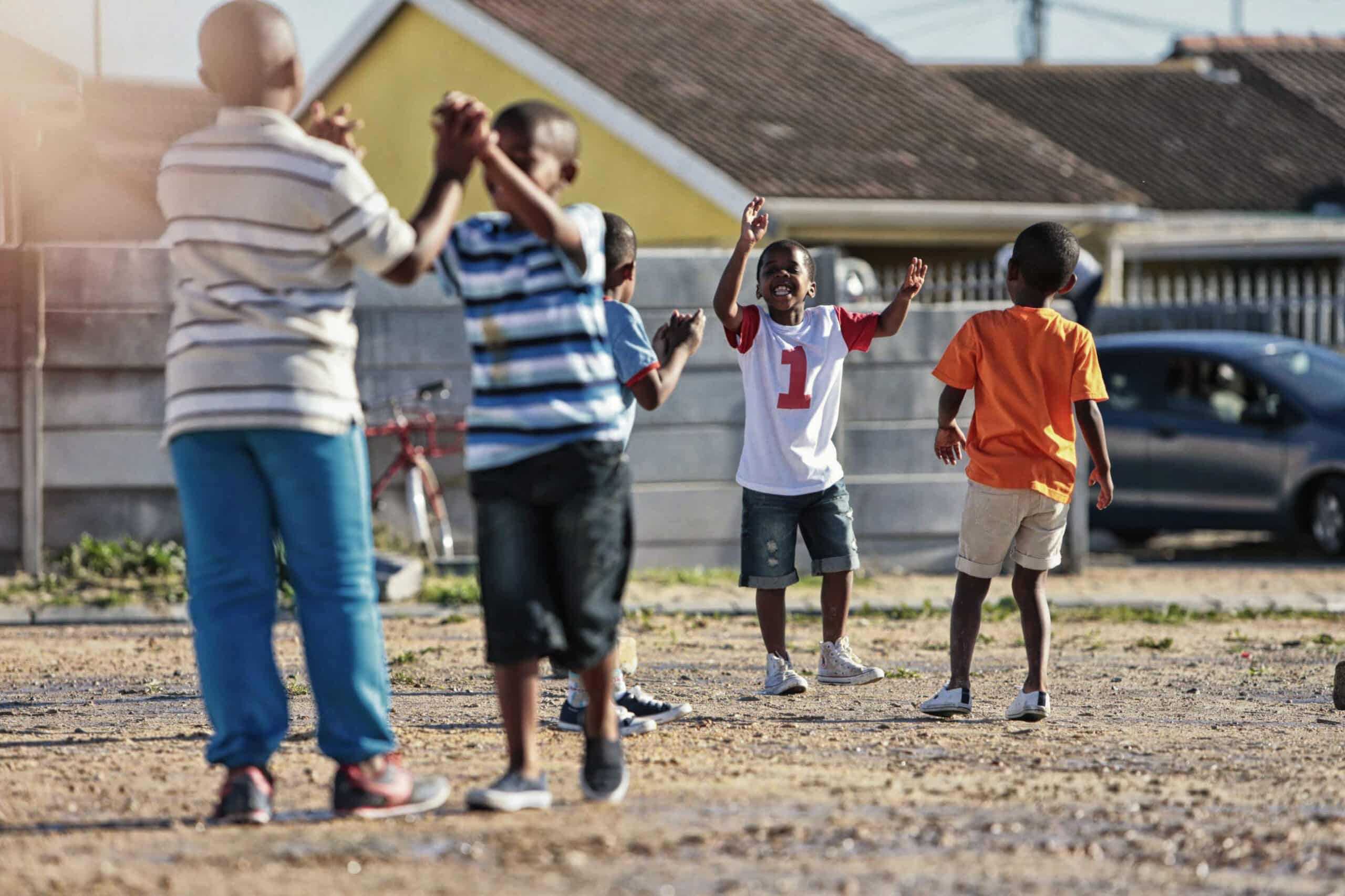 Eine Gruppe Kinder spielt draußen auf einem Feld in einem Wohnviertel zusammen. Ein Kind in einem weiß-roten Hemd steht mit erhobenen Armen da, während die anderen klatschen und rennen. Im Hintergrund sind Häuser und ein geparktes Auto zu sehen. © Fotografie Tomas Rodriguez
