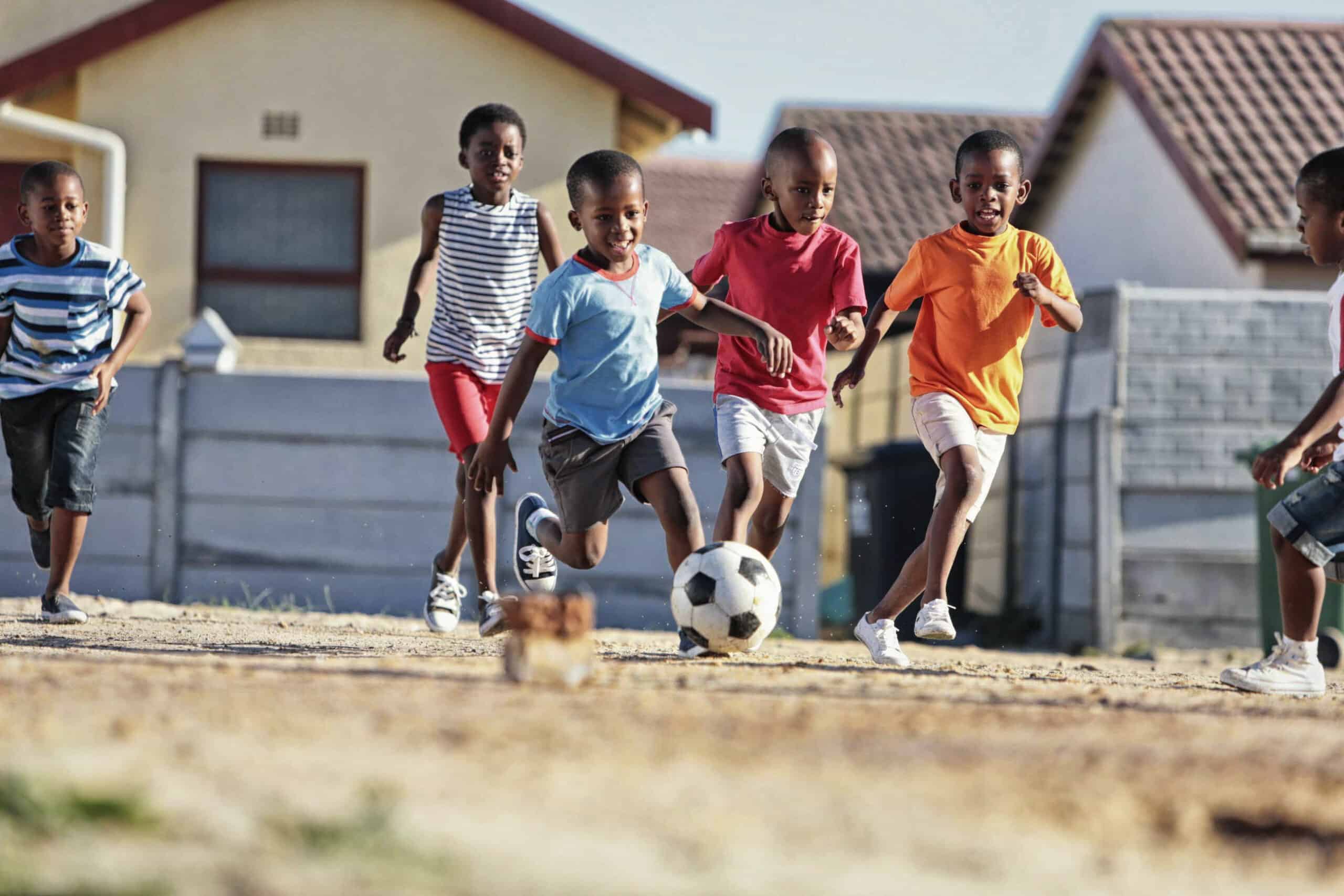 Mehrere Kinder spielen Fußball auf einem Feld in einem Wohngebiet. Sechs Jungen rennen dem Ball entgegen und wetteifern eifrig darum, ihn zu kicken. Die Häuser im Hintergrund haben Ziegeldächer und der Himmel ist klar, was auf einen sonnigen Tag schließen lässt. © Fotografie Tomas Rodriguez