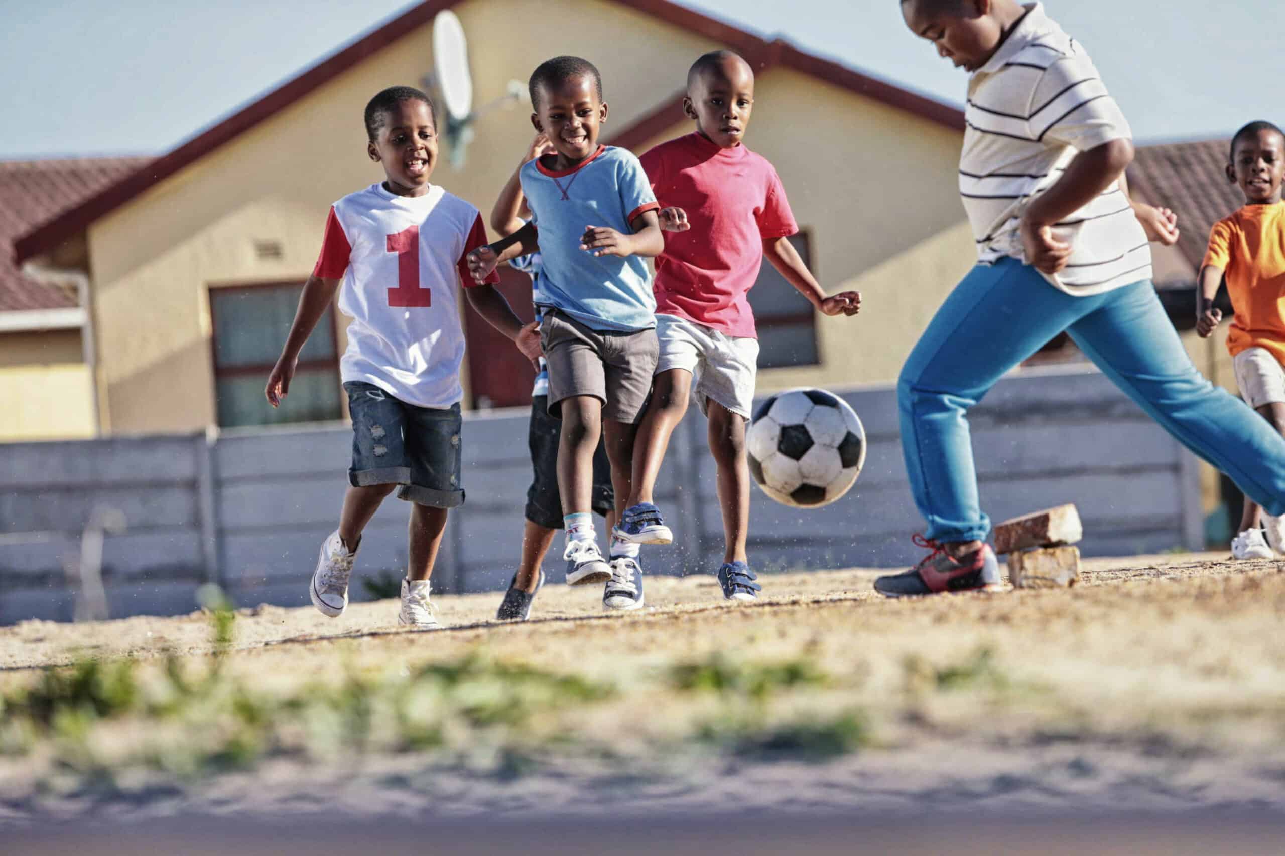 Eine Gruppe junger Jungen spielt an einem sonnigen Tag draußen Fußball. Ein Junge jagt dem Ball hinterher, während die anderen ihm dicht auf den Fersen sind. Sie tragen Freizeitkleidung und im Hintergrund sind Häuser und ein klarer Himmel zu sehen. © Fotografie Tomas Rodriguez