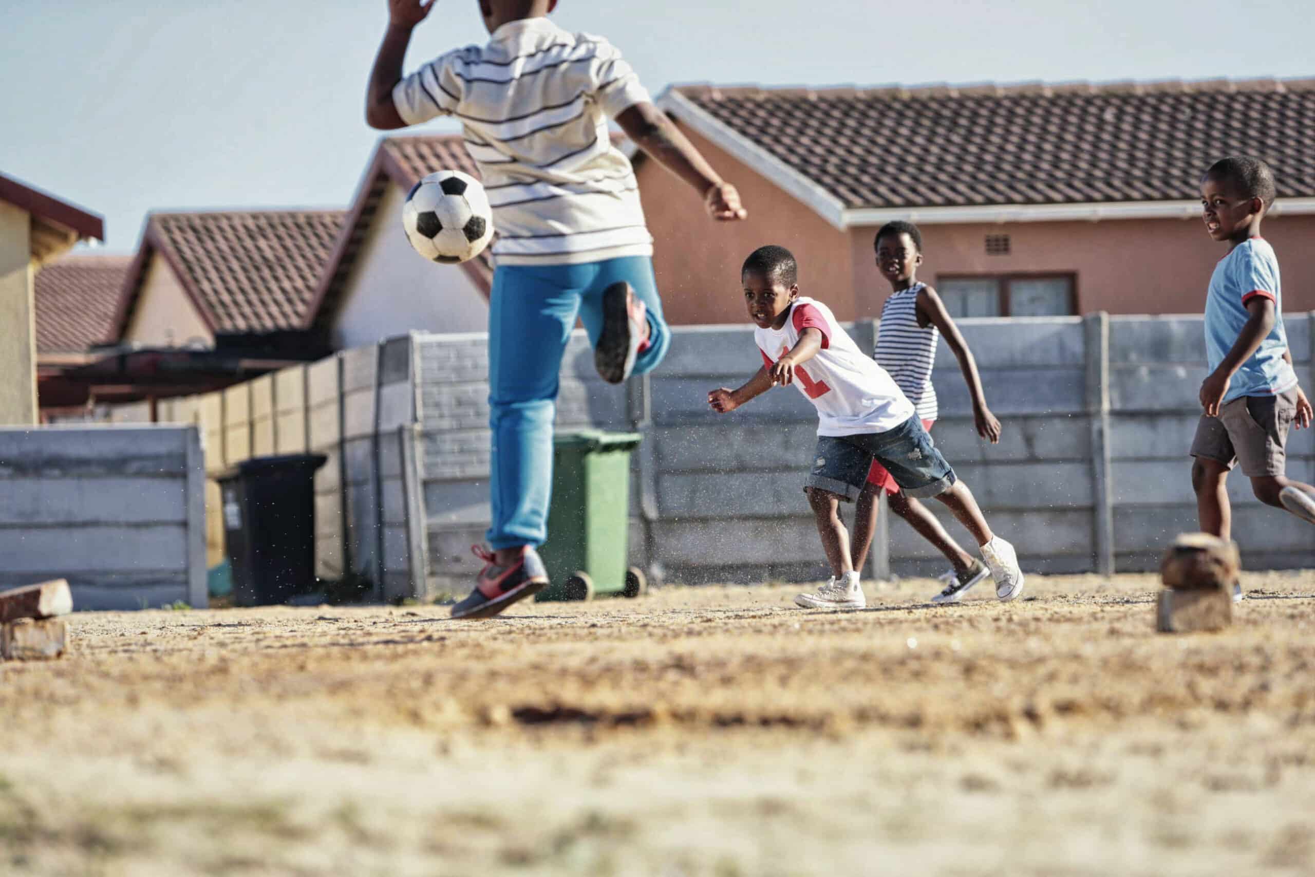 Vier Kinder spielen Fußball auf einem Feld aus Erde, im Hintergrund sind Häuser und ein eingezäunter Bereich zu sehen. Ein Kind kickt den Ball in die Luft, während die anderen drei zuschauen und auf ihn zugehen. Sie sind leger in Shorts und T-Shirts gekleidet. © Fotografie Tomas Rodriguez