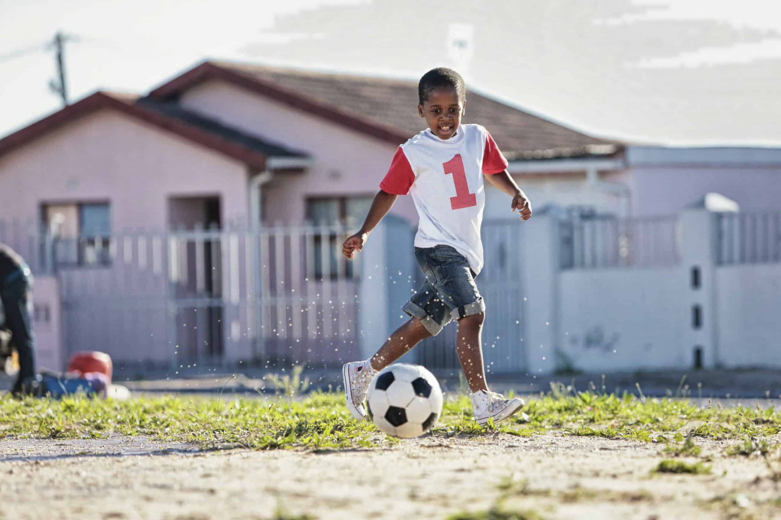 Ein kleiner Junge in einem weiß-roten Trikot mit der Nummer 1 und Jeansshorts kickt einen Fußball auf einer Wiese. Im Hintergrund sind ein rosa Haus und ein weißer Zaun zu sehen, und das Sonnenlicht wirft Schatten um ihn herum. © Fotografie Tomas Rodriguez