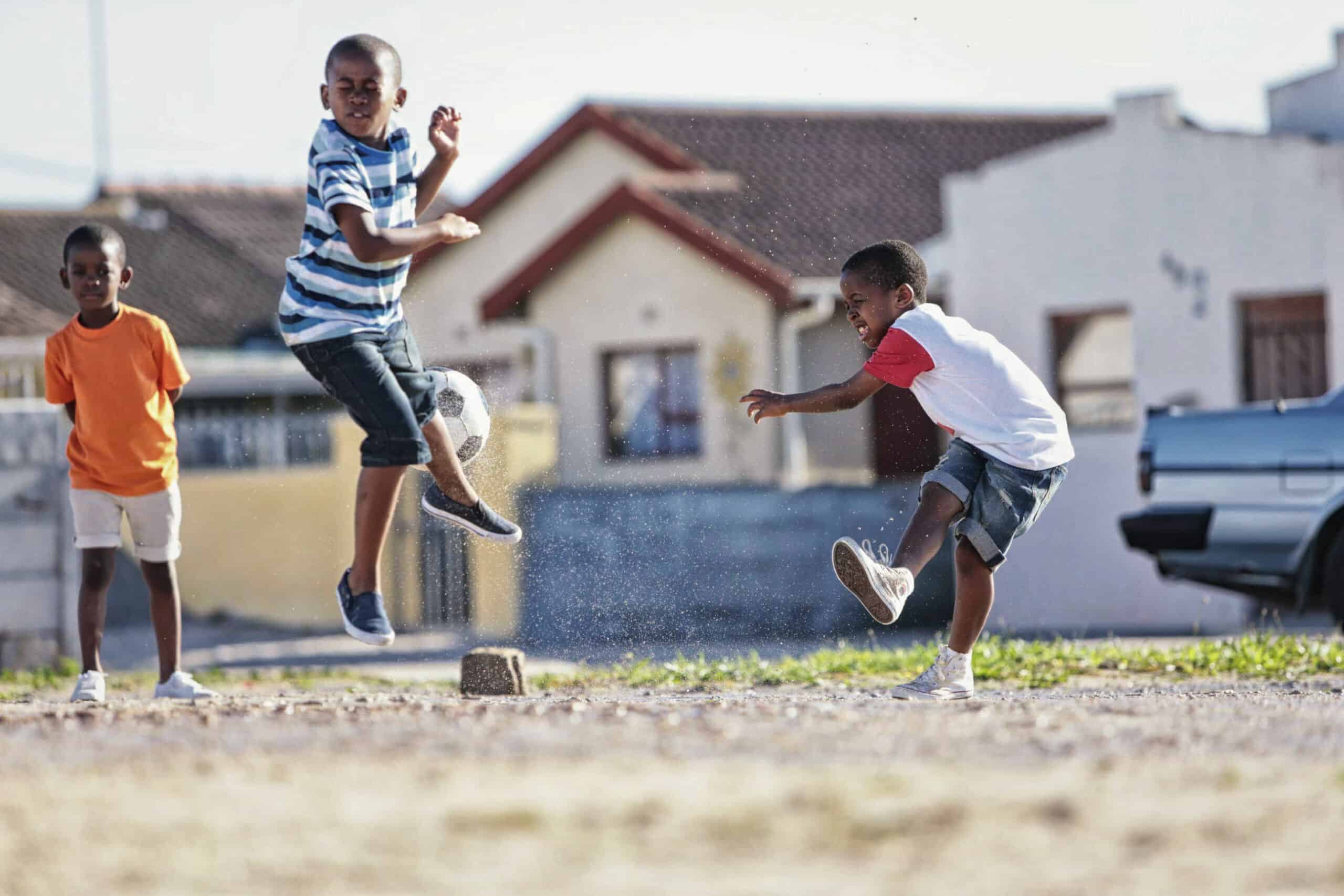 Drei kleine Jungen spielen draußen. Ein Junge in einem orangefarbenen Hemd steht links und schaut zu. In der Mitte springt ein Junge in einem gestreiften Hemd in die Luft. Rechts kickt ein anderer Junge in einem weiß-roten Hemd einen Ball. Sie befinden sich in einem Wohngebiet mit Häusern im Hintergrund. © Fotografie Tomas Rodriguez