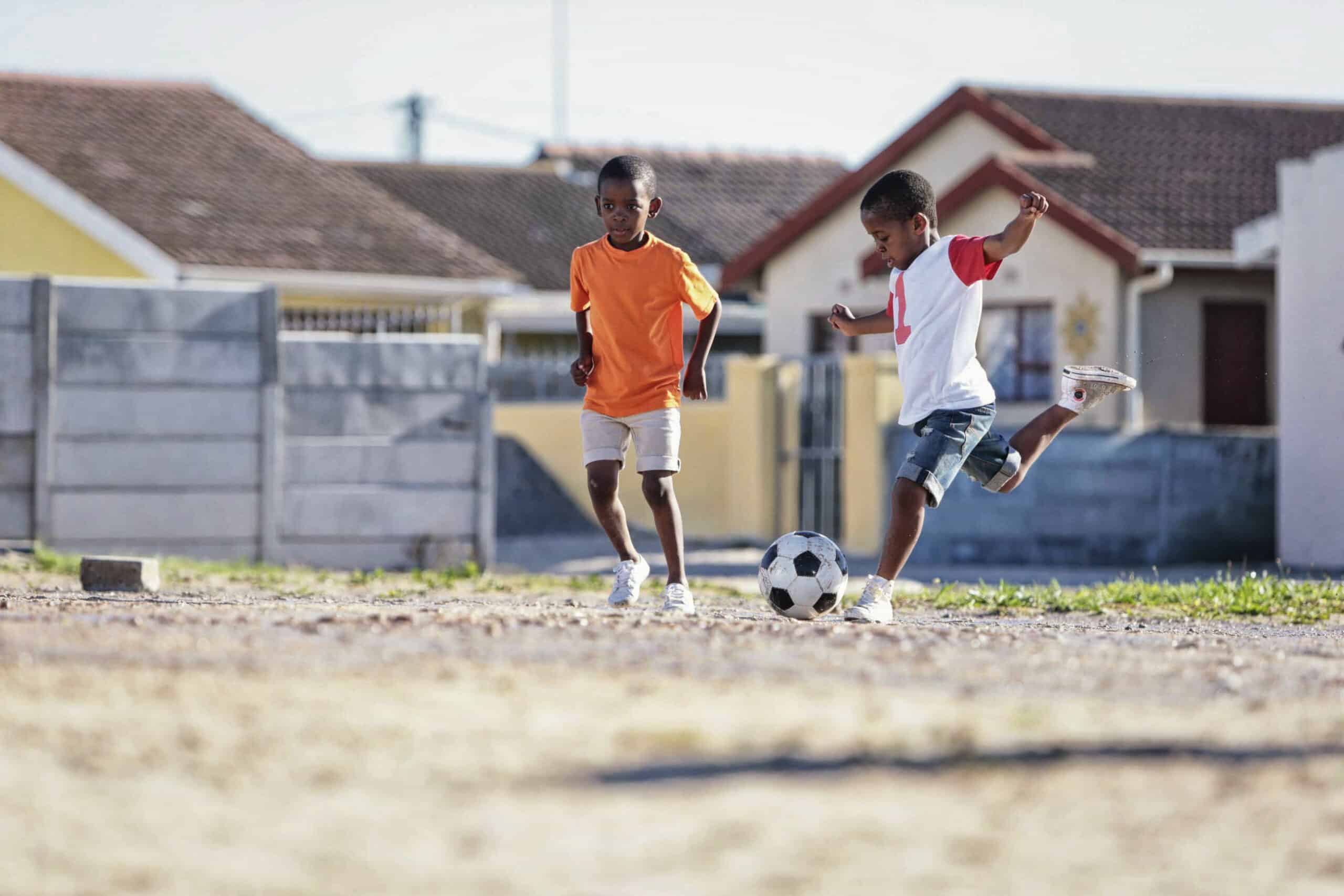 Zwei Jungen spielen an einem sonnigen Tag draußen Fußball. Ein Junge in einem orangefarbenen Hemd rennt, während ein Junge in einem weißen Hemd und Jeansshorts gerade dabei ist, einen schwarz-weißen Fußball zu kicken. Sie befinden sich in einem Viertel mit Häusern und einer Mauer im Hintergrund. © Fotografie Tomas Rodriguez