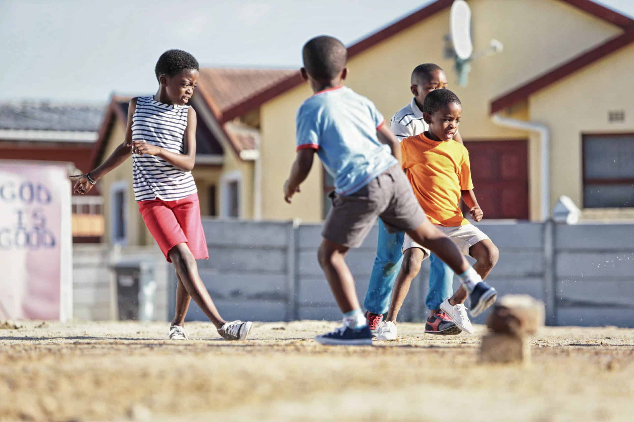 Vier Kinder spielen Fußball auf einem Feld vor Häusern. Eines kickt den Ball, während die anderen herumrennen. Im Hintergrund sind Häuser mit einer Satellitenschüssel zu sehen, und es gibt ein Schild, auf dem teilweise „Gott ist gut“ steht. © Fotografie Tomas Rodriguez
