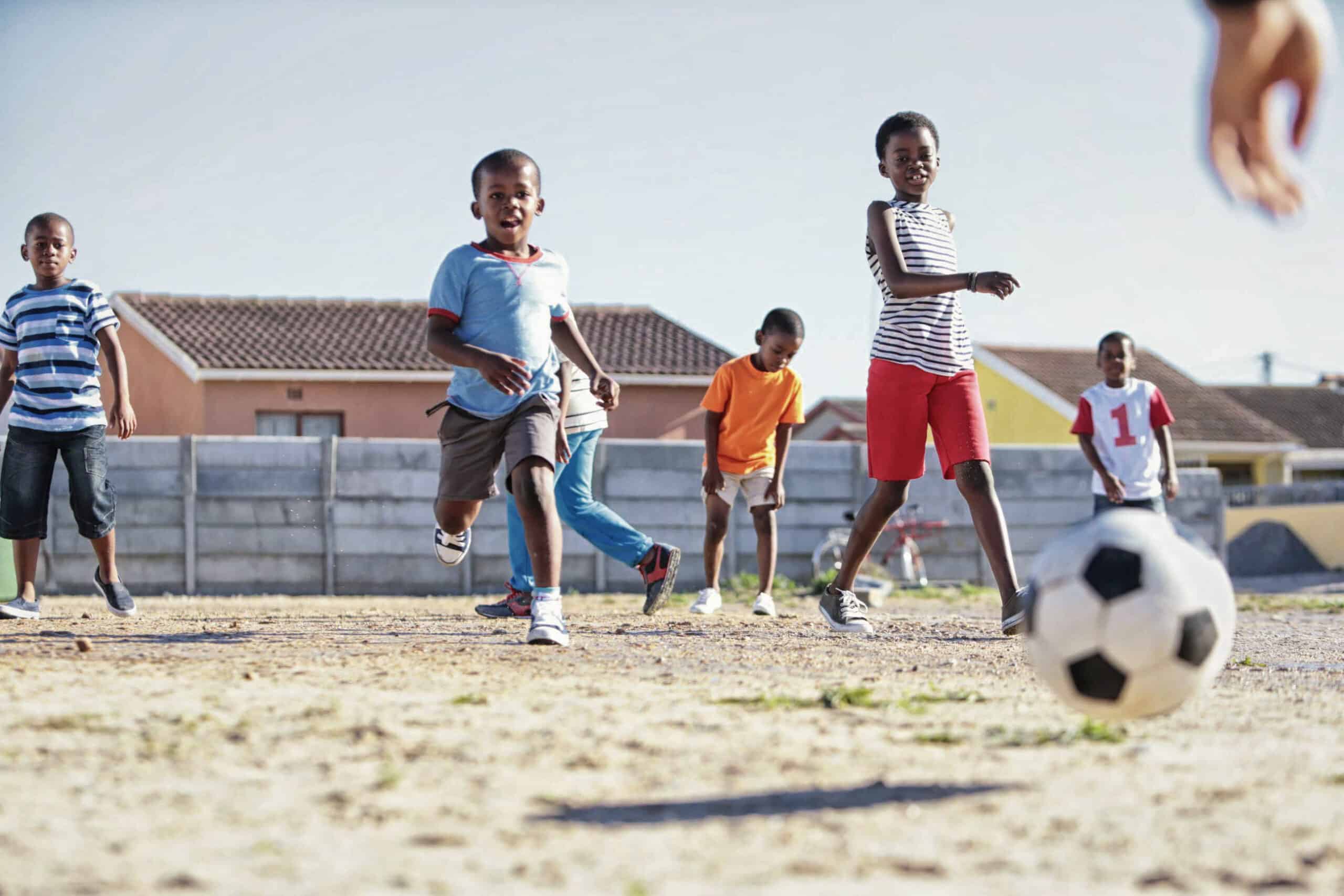 Eine Gruppe Kinder spielt Fußball auf einem Sandplatz. Der Junge vorne kickt den Ball, während die anderen lächelnd und lachend darauf zulaufen. Im Hintergrund sind Häuser mit Ziegeldächern unter einem klaren blauen Himmel zu sehen. © Fotografie Tomas Rodriguez
