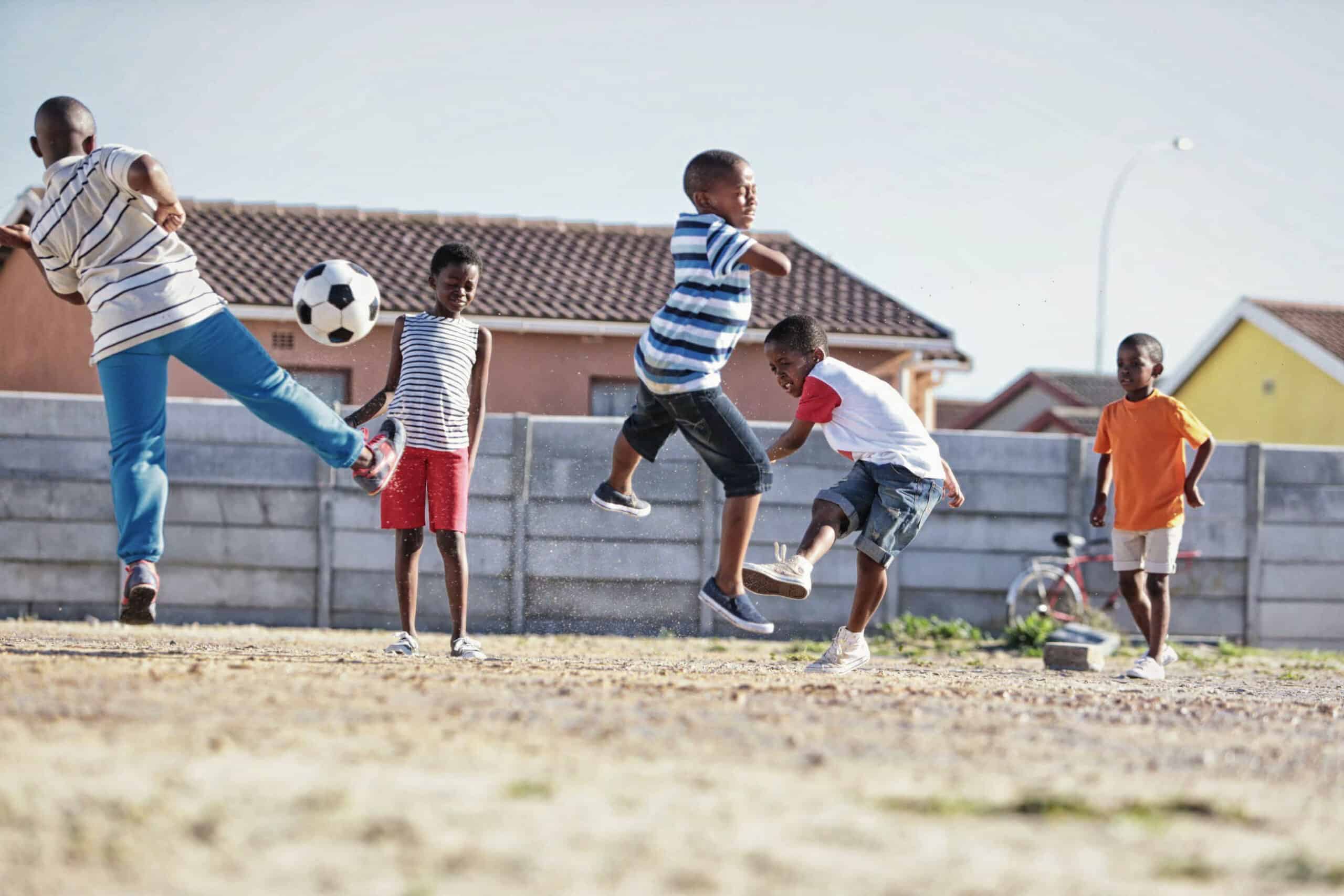 Eine Gruppe Kinder spielt Fußball auf einem Feld in der Nähe eines Wohngebiets. Ein Kind ist in der Luft und will gerade den Ball kicken, während die anderen zuschauen oder mitspielen. Im Hintergrund sind Häuser und ein Fahrrad zu sehen. Das Wetter scheint sonnig und klar. © Fotografie Tomas Rodriguez