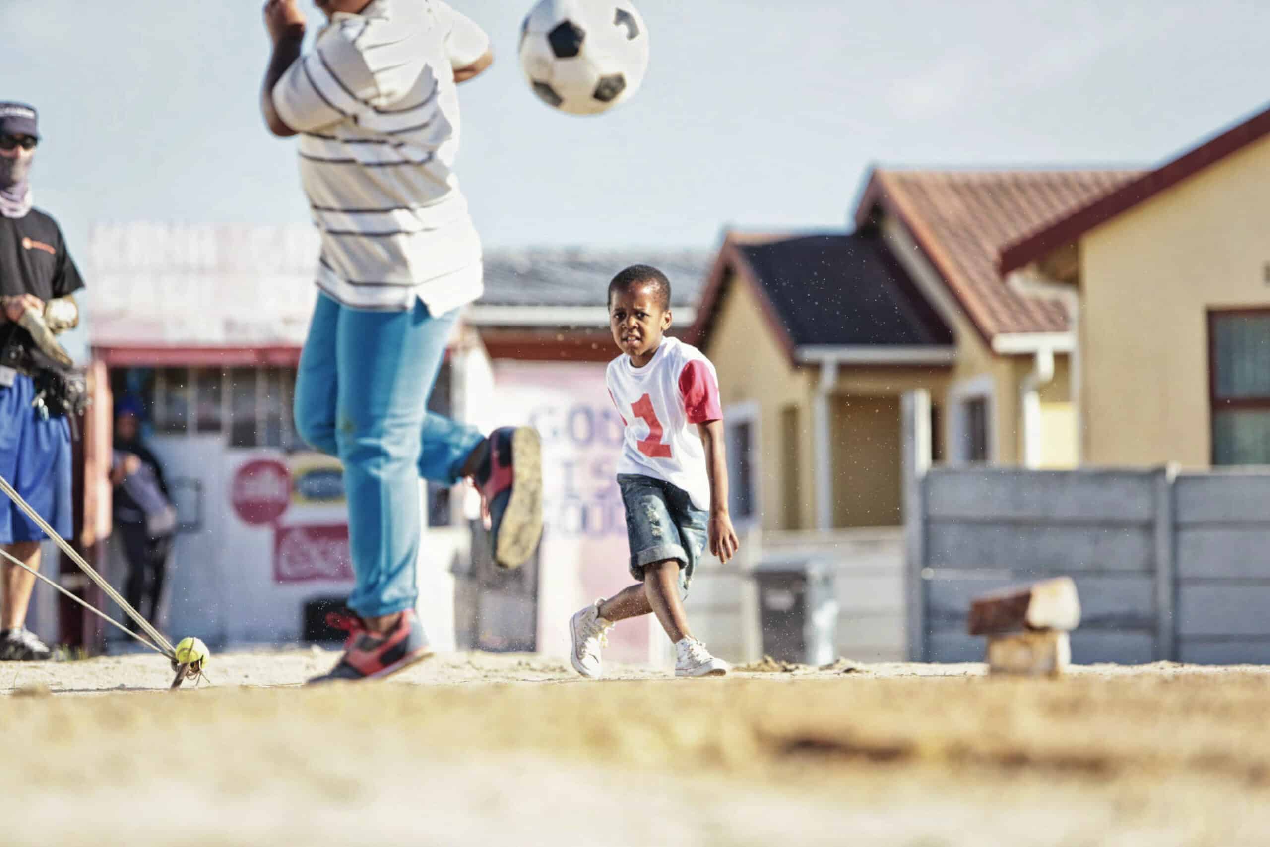 Ein kleiner Junge in einem weiß-roten Hemd beobachtet einen Fußball in der Luft, während ein anderes Kind, das größtenteils außerhalb des Bildes liegt, den Ball kickt. Sie spielen auf einem Feld aus Erde, im Hintergrund sind Häuser und andere Menschen zu sehen. Der Himmel ist klar, was auf einen sonnigen Tag schließen lässt. © Fotografie Tomas Rodriguez