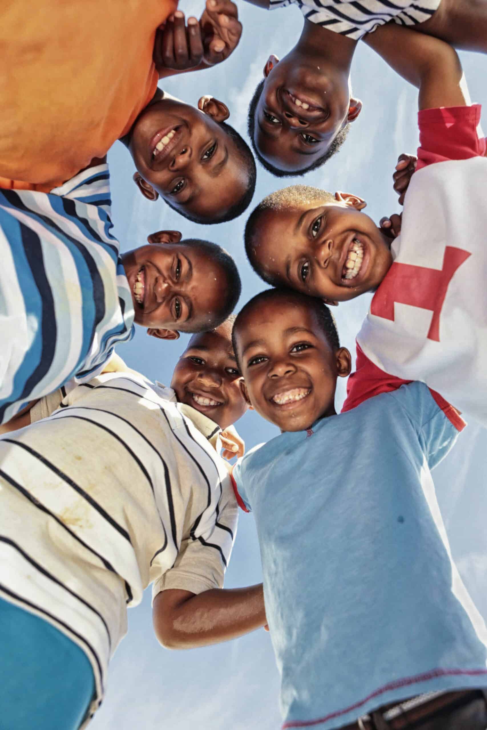 Eine Gruppe von sechs kleinen Kindern steht dicht beieinander in einem Kreis, lächelt strahlend und blickt in die Kamera. Sie sind an einem sonnigen Tag im Freien vor einem blauen Himmel. Jedes Kind ist farbenfroh gekleidet und strahlt Freude und Kameradschaft aus. © Fotografie Tomas Rodriguez