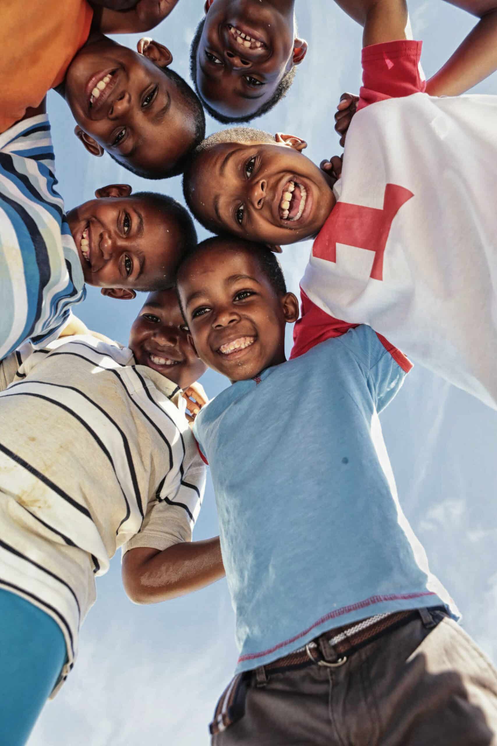 Eine Gruppe von sechs Kindern drängt sich freudig in einem Kreis zusammen, die Köpfe eng beieinander, lächeln breit und blicken in die Kamera. Der Himmel dient als heller und klarer Hintergrund. Die Ausdrücke der Kinder vermitteln ein Gefühl von Glück und Kameradschaft. © Fotografie Tomas Rodriguez