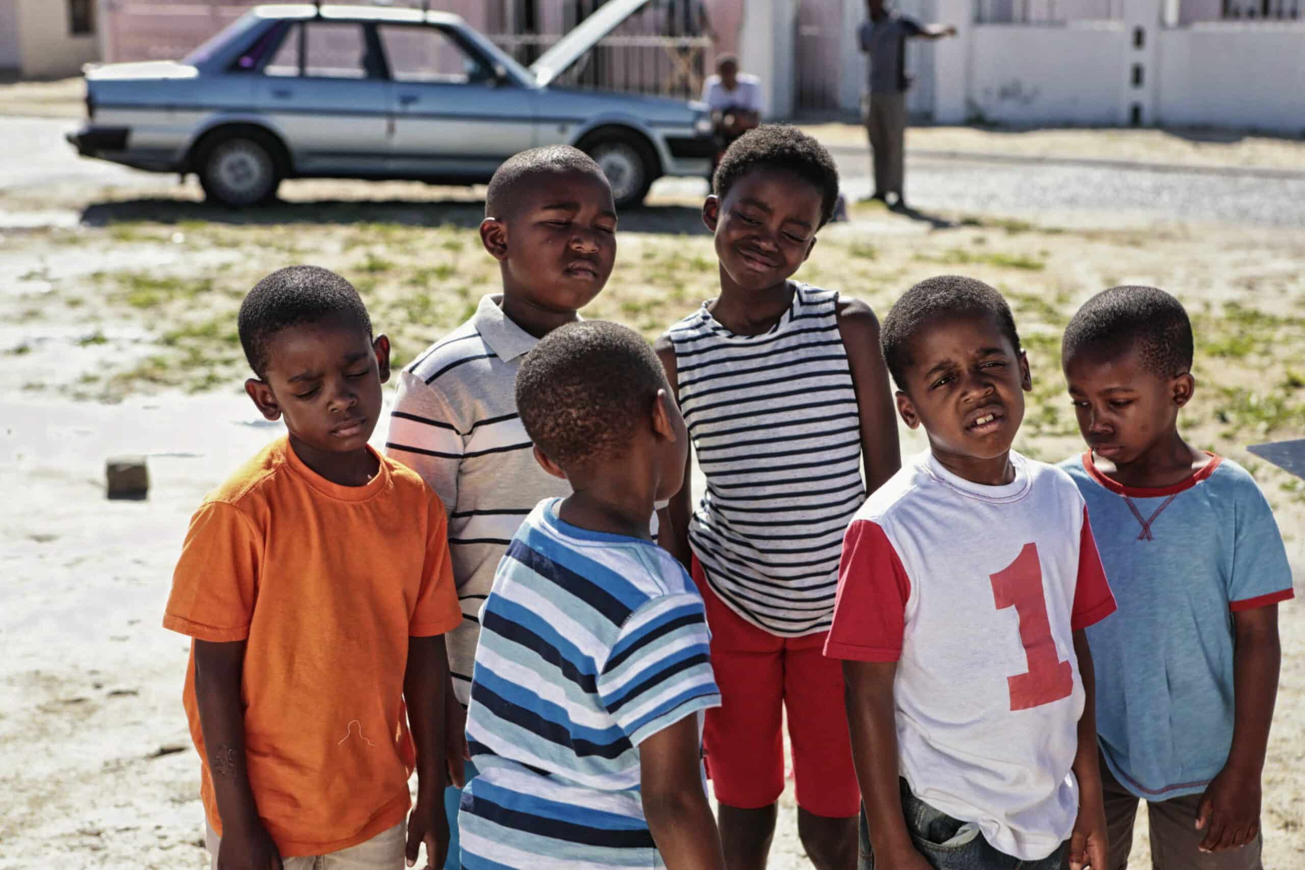 Eine Gruppe von sechs Kindern steht an einem sonnigen Tag draußen zusammen. Sie tragen Freizeitkleidung und bunte Hemden. Im Hintergrund sind ein geparktes Auto und ein paar Gebäude zu sehen. Einige der Kinder haben wegen des Sonnenlichts die Augen geschlossen oder blinzeln. © Fotografie Tomas Rodriguez