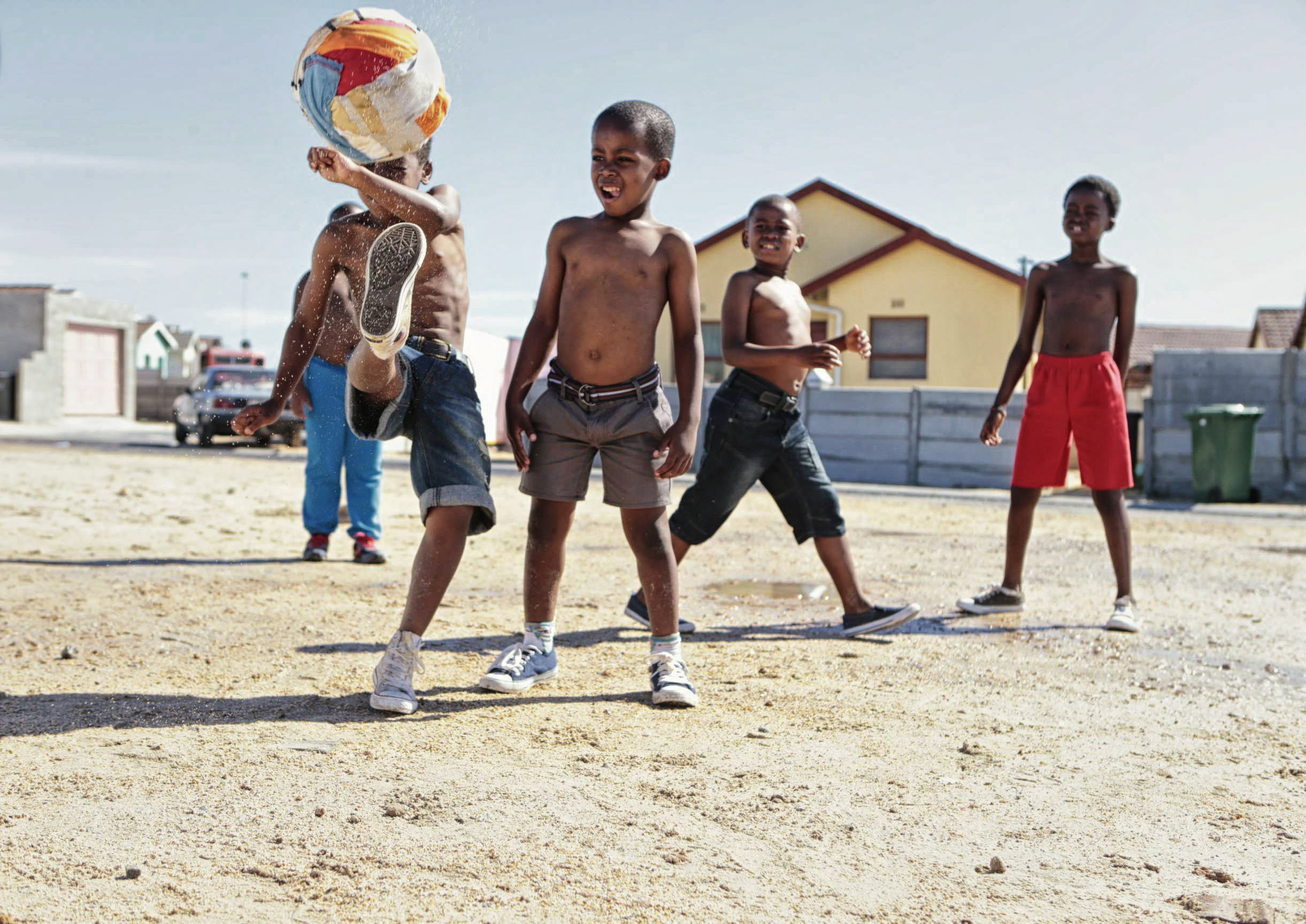 Eine Gruppe junger Jungen spielt auf einem Sandplatz zwischen Häusern Fußball. Ein Junge, ohne Hemd und in grauen Shorts, kickt einen bunten Fußball, während die anderen lächelnd zuschauen. Die Szene ist hell und sonnig, mit einstöckigen Häusern und ein paar anderen Kindern im Hintergrund. © Fotografie Tomas Rodriguez