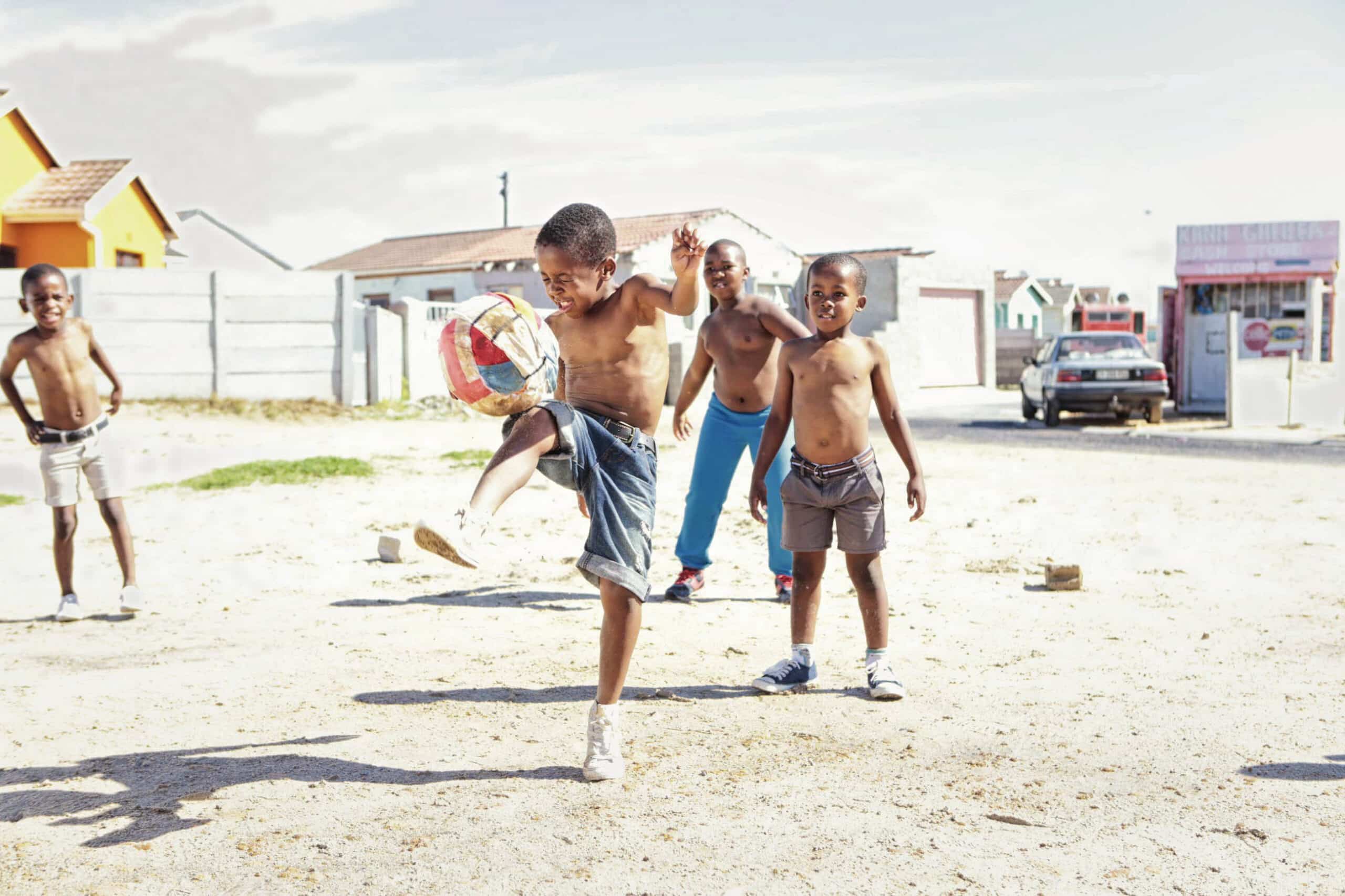 Fünf junge Jungen, alle mit nacktem Oberkörper, spielen auf einem offenen Sandplatz Fußball. Ein Junge kickt zielstrebig einen rot-weißen Ball, während die anderen ihm zuschauen und ihn anfeuern. Im Hintergrund sind Häuser und ein geparktes Auto zu sehen. Die Atmosphäre ist lebhaft und energiegeladen. © Fotografie Tomas Rodriguez