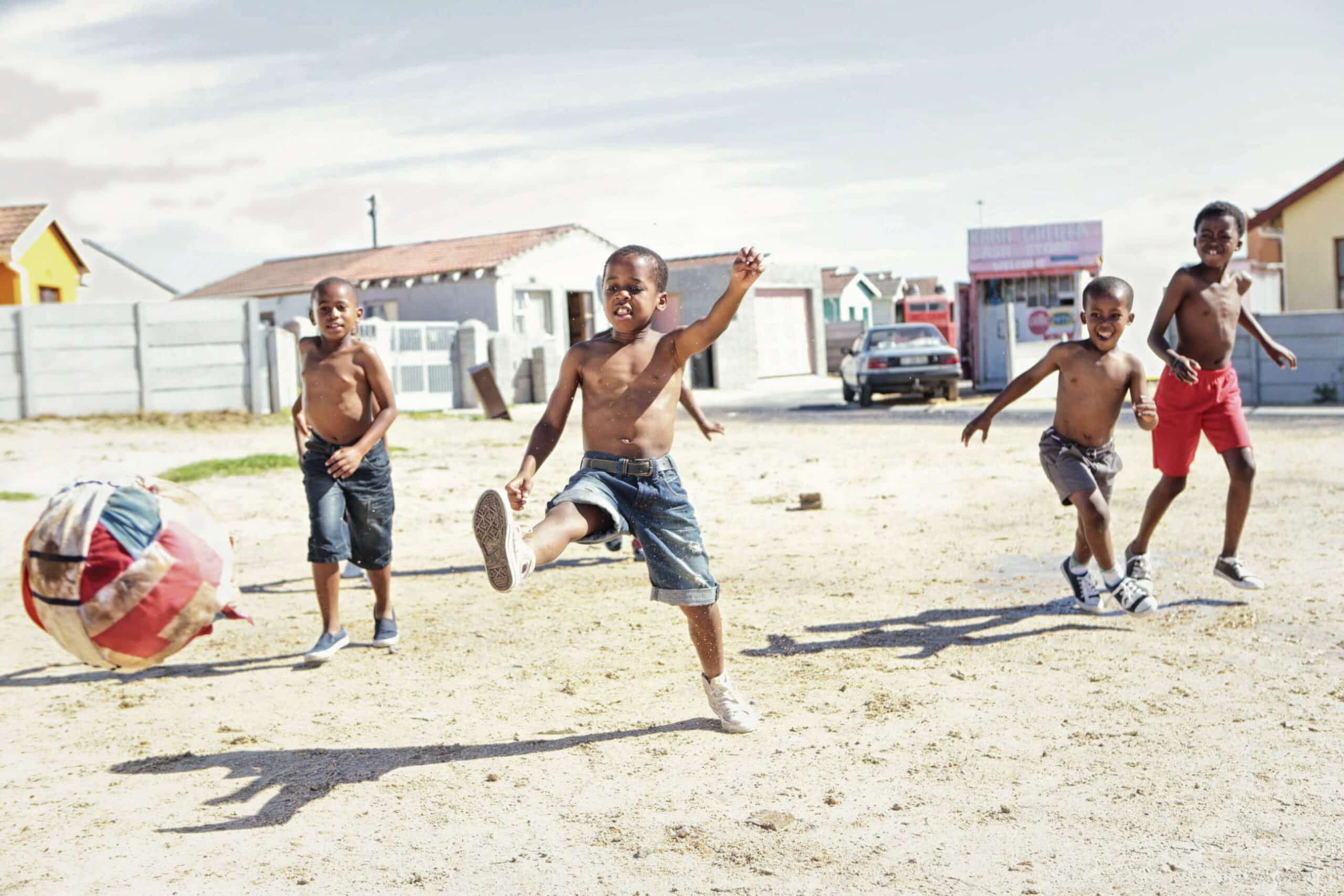 Vier Jungen spielen auf einem Sandspielplatz mit einem bunten Ball Fußball. Ein Junge, ohne Hemd und in Jeansshorts, kickt den Ball, während die anderen, ebenfalls ohne Hemd, herumlaufen und zuschauen. Im Hintergrund sind Häuser und ein Auto zu sehen. © Fotografie Tomas Rodriguez