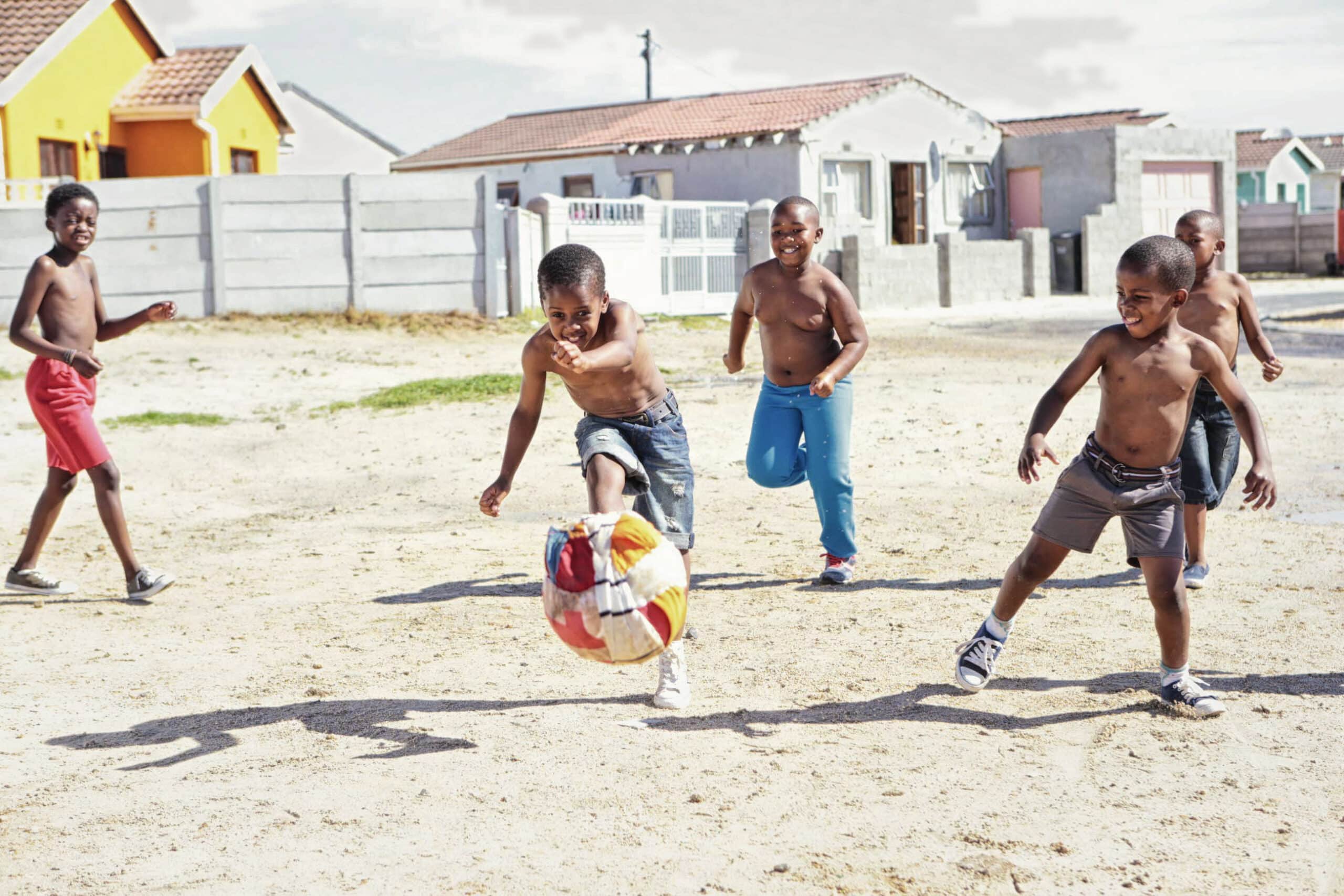 Fünf Jungen spielen mit nacktem Oberkörper Fußball auf einem Sandplatz in einem Wohngebiet mit bunten Häusern im Hintergrund. Ein Junge kickt einen großen bunten Ball, während die anderen rennen und lachen und das Spiel unter einem klaren Himmel genießen. © Fotografie Tomas Rodriguez