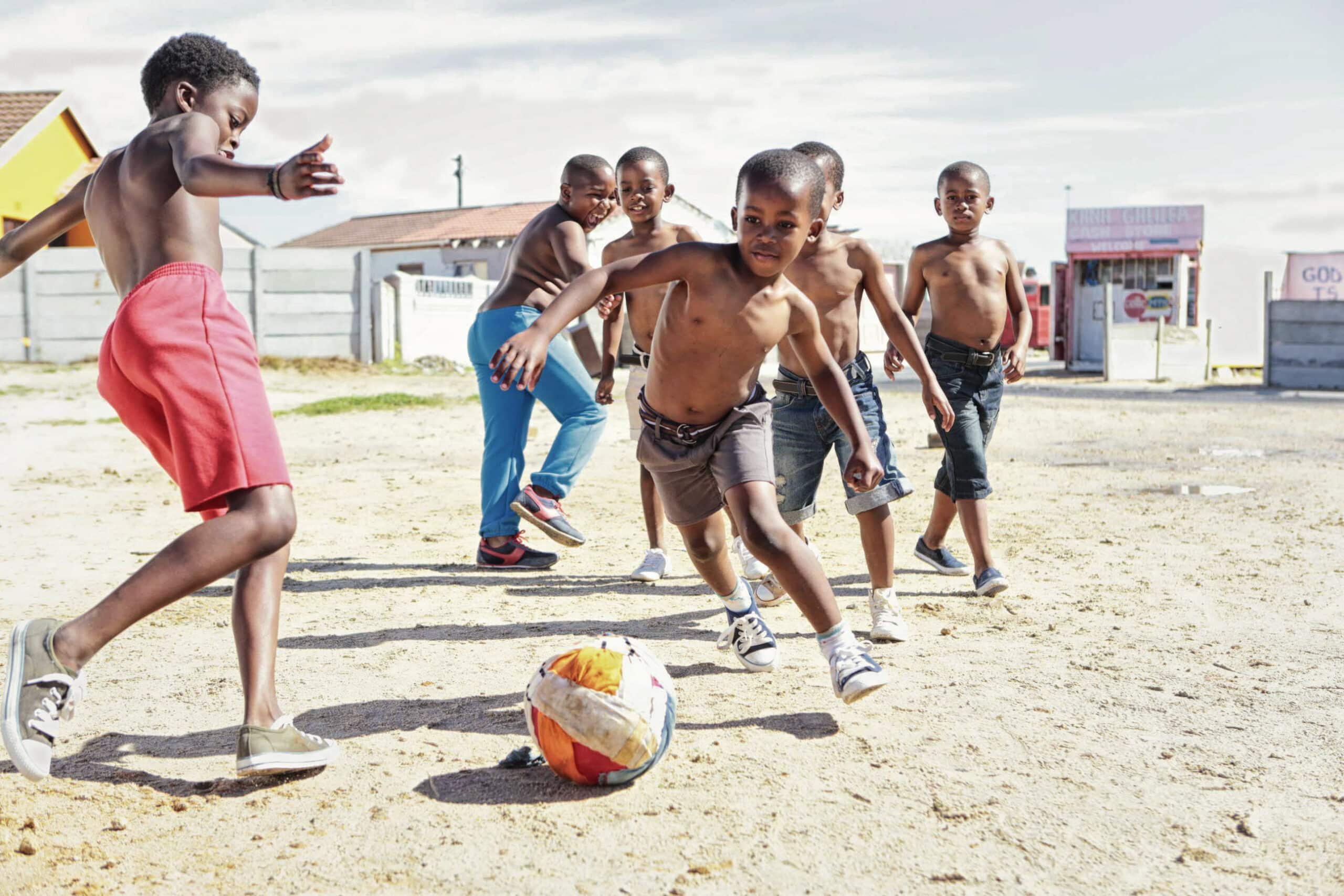 Eine Gruppe von Jungen spielt Fußball auf einem Sandplatz. Sie sind oben ohne und scheinen voller Begeisterung einem bunten, abgenutzten Fußball hinterherzujagen und ihn zu kicken. Im Hintergrund sind Häuser, ein Zaun und ein entferntes Gebäude mit einem Schild zu sehen. © Fotografie Tomas Rodriguez