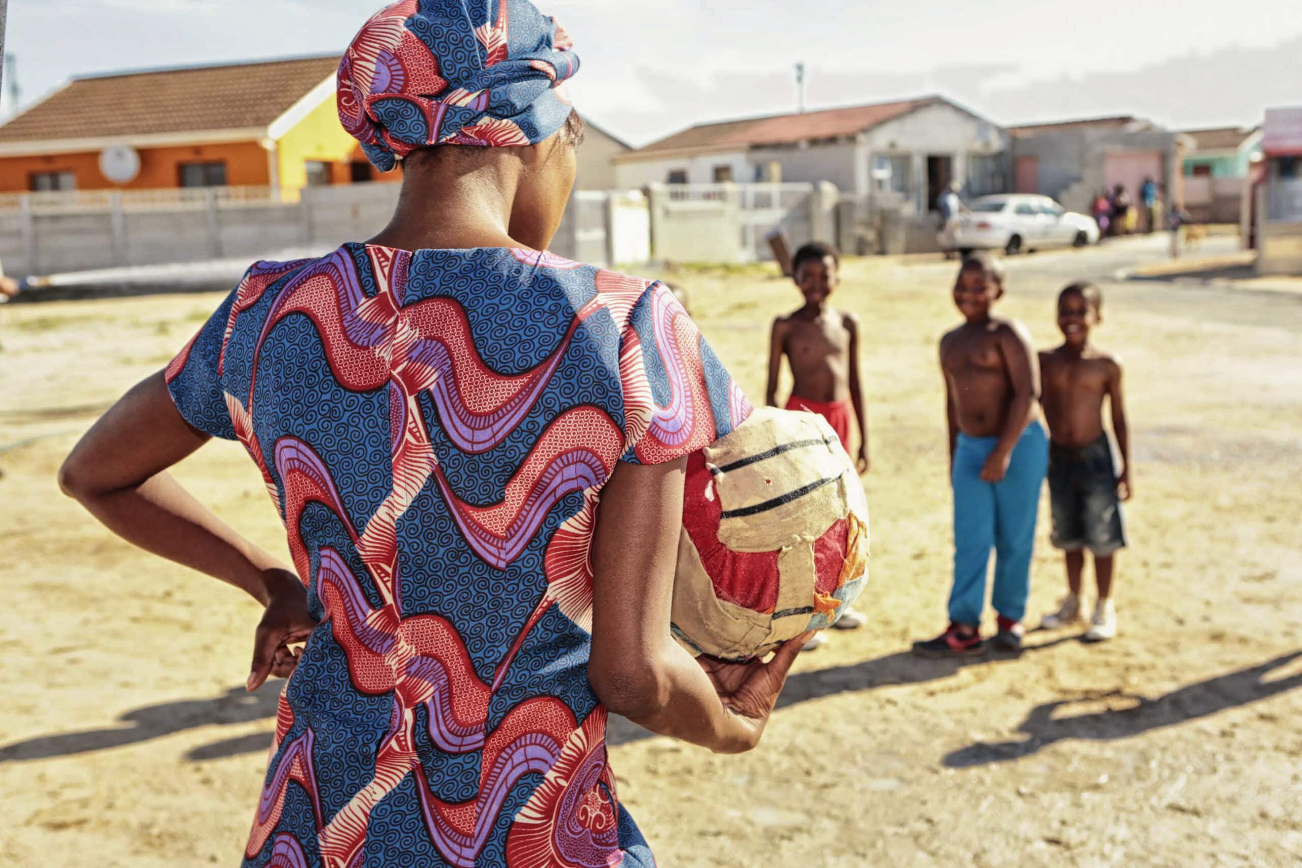 Eine Frau in einem bunten Kleid und Kopftuch steht mit dem Rücken zur Kamera und hält einen selbstgemachten Fußball in der Hand. Drei Jungen ohne Hemd stehen lächelnd vor ihr, im Hintergrund sind Häuser und ein Auto an einem sonnigen Tag zu sehen. © Fotografie Tomas Rodriguez