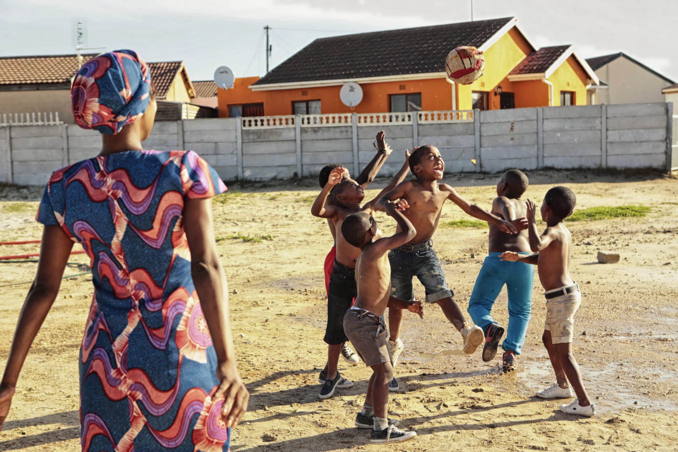 Eine Frau in einem bunten Kleid steht da und beobachtet eine Gruppe von Kindern ohne Hemd, die in einem sandigen Hof mit einem Ball spielen. Im Hintergrund sind Häuser mit orangefarbenen und beigen Wänden und Satellitenschüsseln zu sehen. Eine Betonmauer trennt den Hof von den Häusern. © Fotografie Tomas Rodriguez