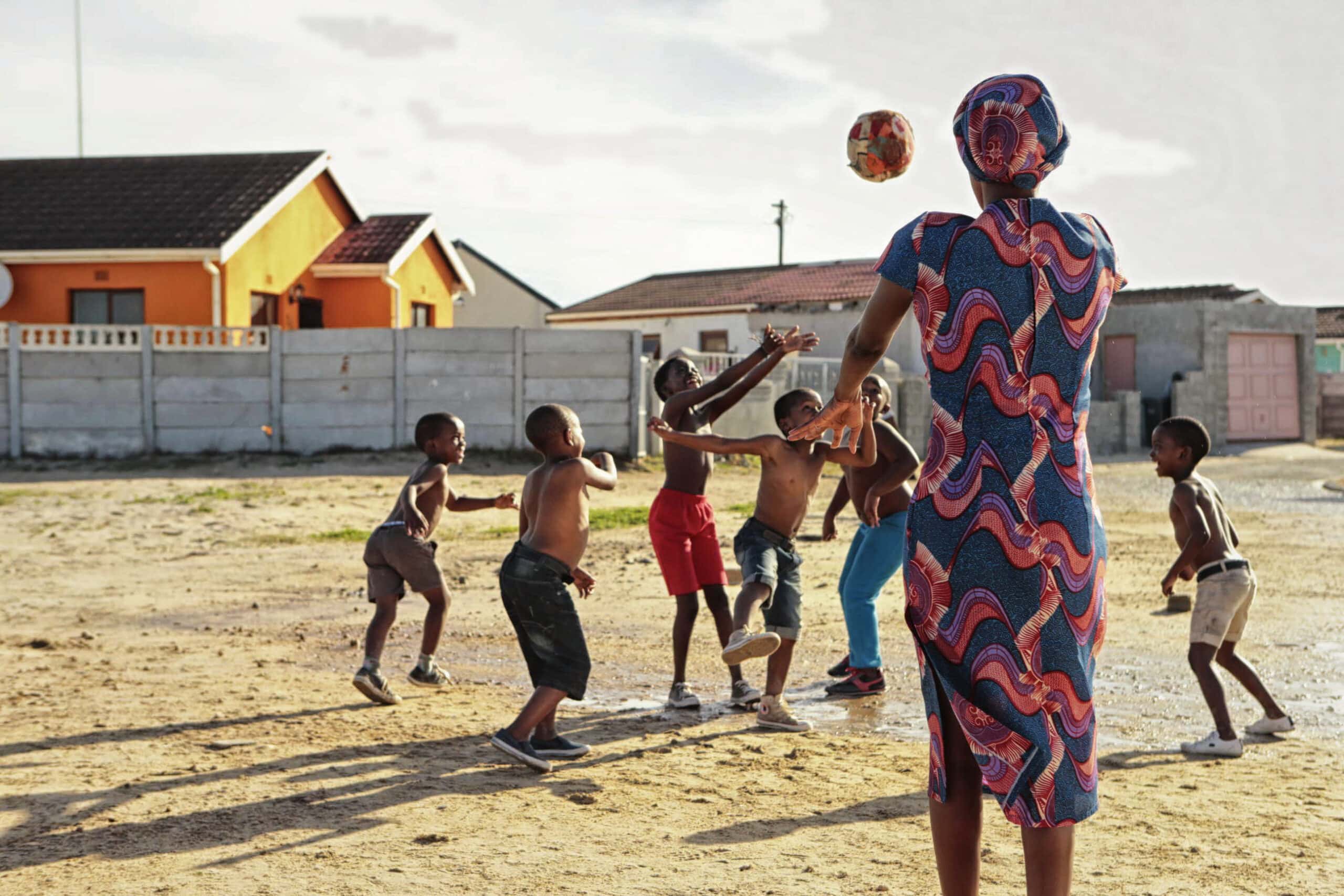 Eine Frau in einem bunten Kleid spielt mit einer Gruppe von Kindern auf einem offenen, sandigen Gelände. Sie wirft ihnen einen Ball zu, und die Kinder greifen eifrig nach ihm, um ihn zu fangen. Im Hintergrund sind einstöckige Häuser mit orangefarbenen Außenfassaden und einer grauen Wand zu sehen. © Fotografie Tomas Rodriguez