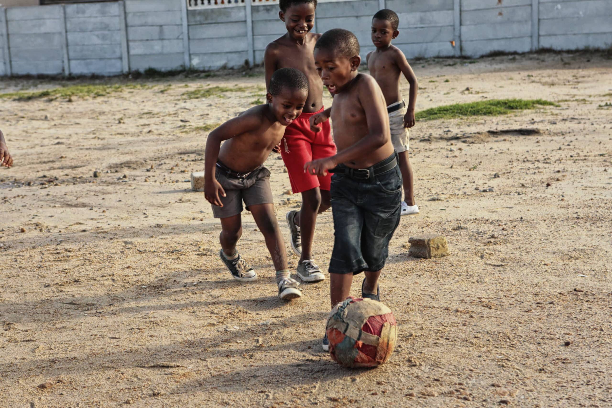 Vier junge Jungen, mit nacktem Oberkörper und barfuß, spielen Fußball auf einem Sandplatz. Ein Junge geht mit dem Ball voran, während ein anderer lächelnd hinter ihm herläuft. Zwei weitere Jungen im Hintergrund schauen zu und laufen nebenher. In der Ferne ist eine Betonmauer zu sehen. © Fotografie Tomas Rodriguez