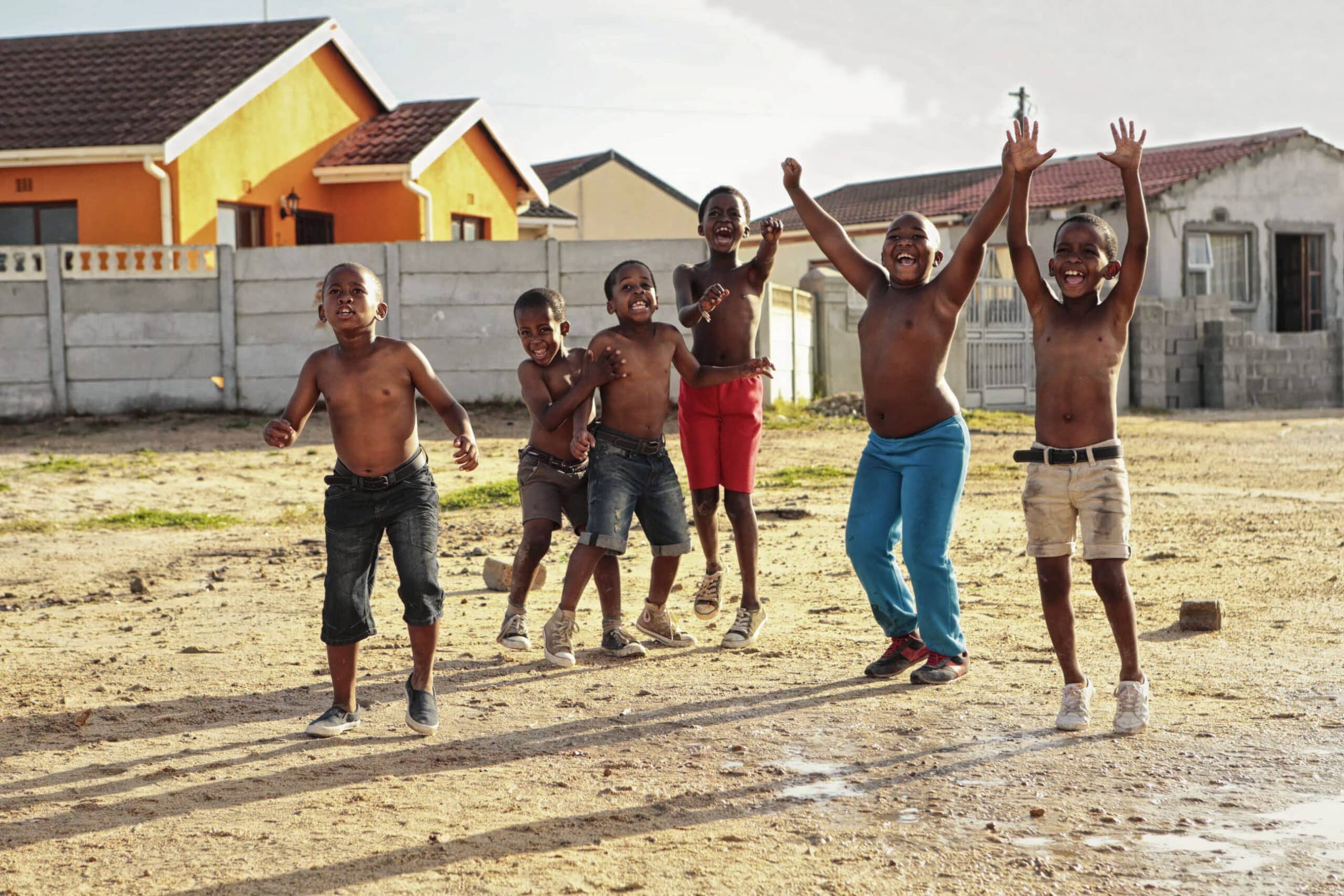 Eine Gruppe von sechs glücklichen Kindern spielt an einem sonnigen Tag draußen zusammen. Sie sind alle oben ohne, bis auf eines in einer blauen Hose. Einige klatschen und springen vor Freude auf einem Sandboden mit Häusern im Hintergrund, was eine lebhafte und fröhliche Szene schafft. © Fotografie Tomas Rodriguez