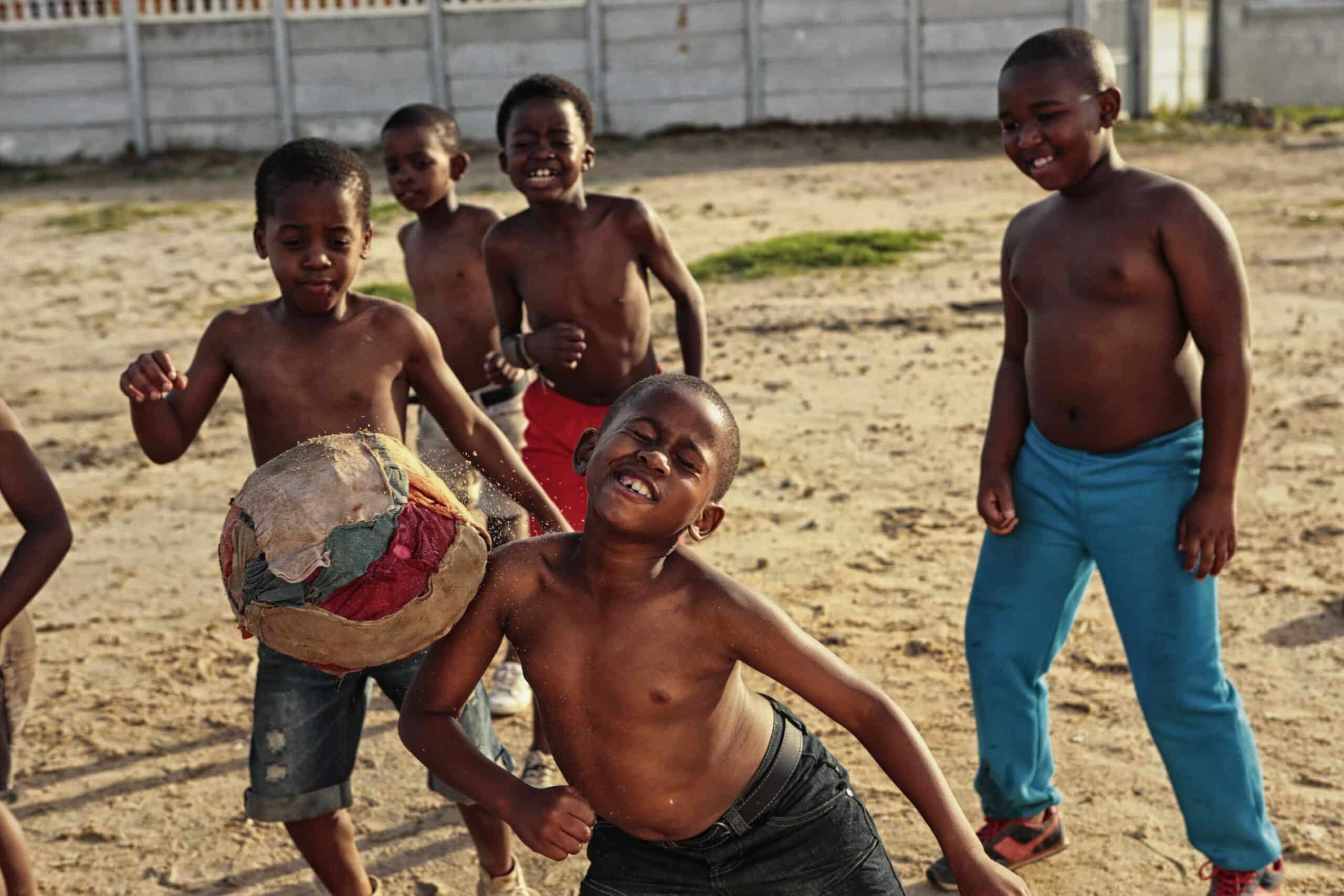 Eine Gruppe von sechs Jungen spielt mit einem abgenutzten Fußball auf einem Feld aus Erde. Sie sind alle oben ohne, manche tragen Shorts, während einer blaue Hosen trägt. Der Junge im Vordergrund ist gerade dabei, den Ball zu kicken. Sie lächeln und scheinen Spaß zu haben. © Fotografie Tomas Rodriguez