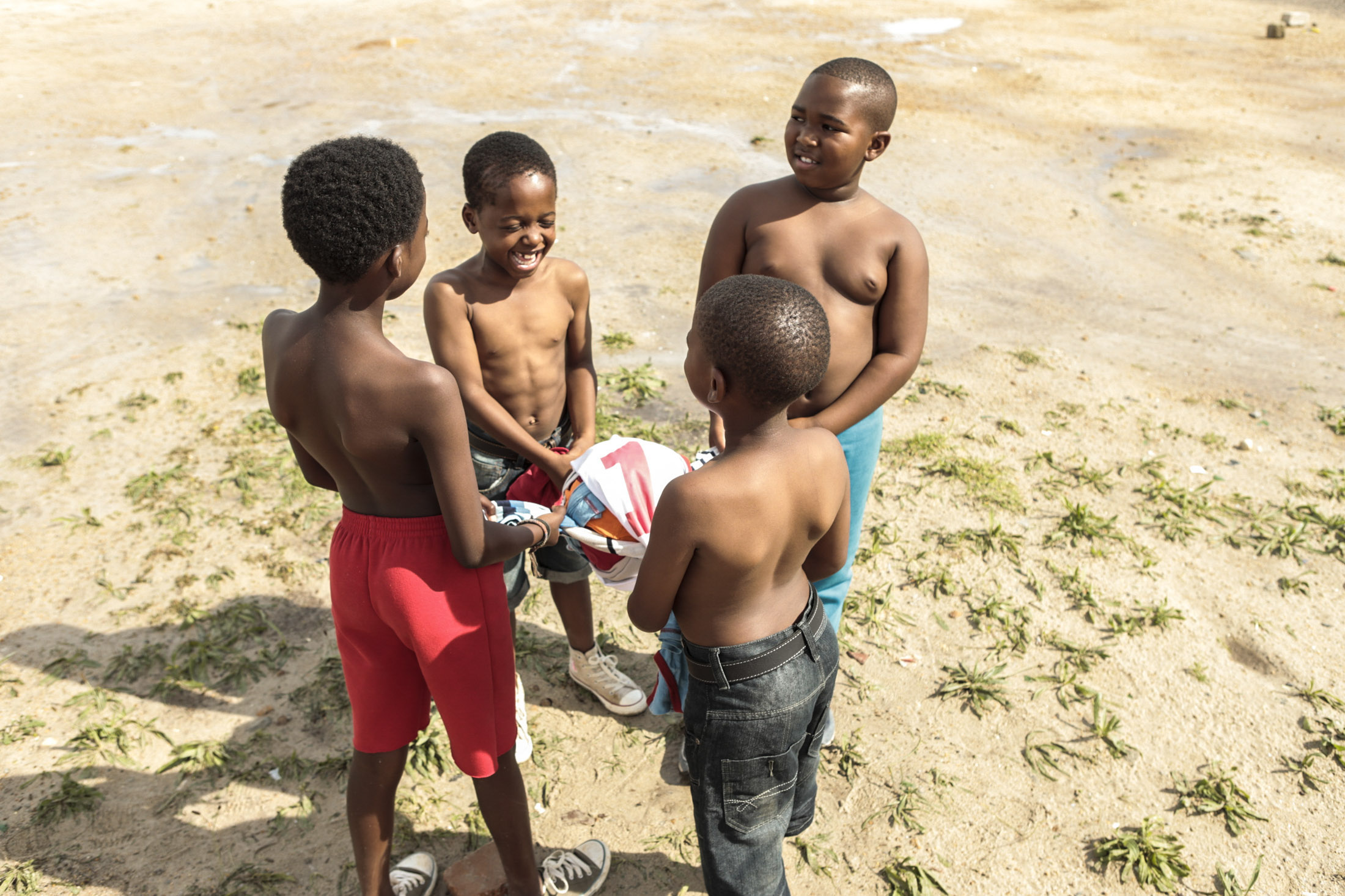 Vier oberkörperfreie Jungen stehen in einem Kreis auf einer offenen, sandigen Fläche und halten einen bunten Fußball in den Händen. Sie scheinen zu lachen und ihre gemeinsame Zeit zu genießen. Auf dem Boden sind vereinzelt grüne Grasflecken zu sehen. © Fotografie Tomas Rodriguez