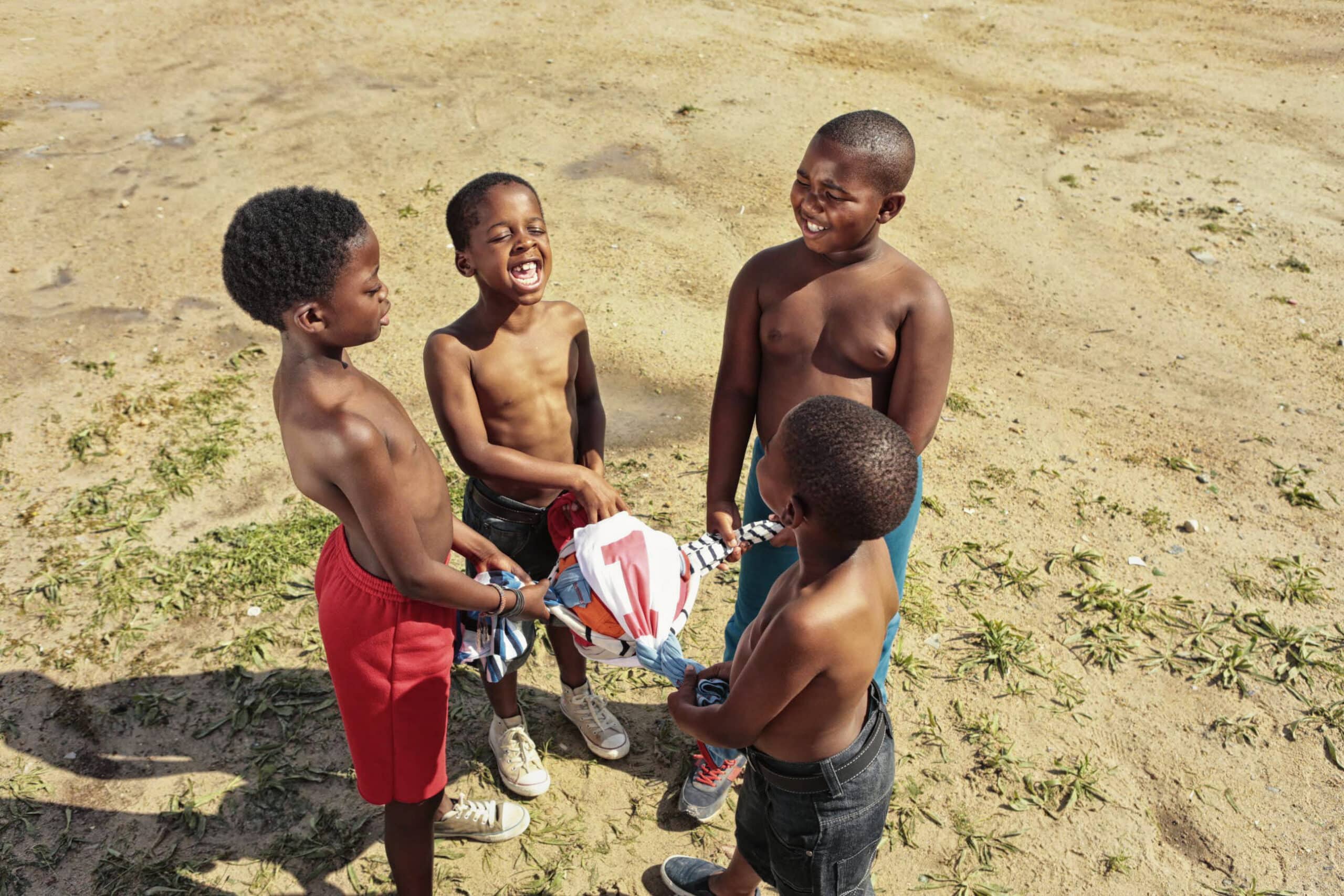 Vier Kinder mit kurzen Haaren und ohne Hemd stehen auf staubigem Boden, halten ein Stoffbündel und lächeln und lachen gemeinsam. Die Szene strahlt ein Gefühl von Freude und Kameradschaft unter den Kindern aus. © Fotografie Tomas Rodriguez
