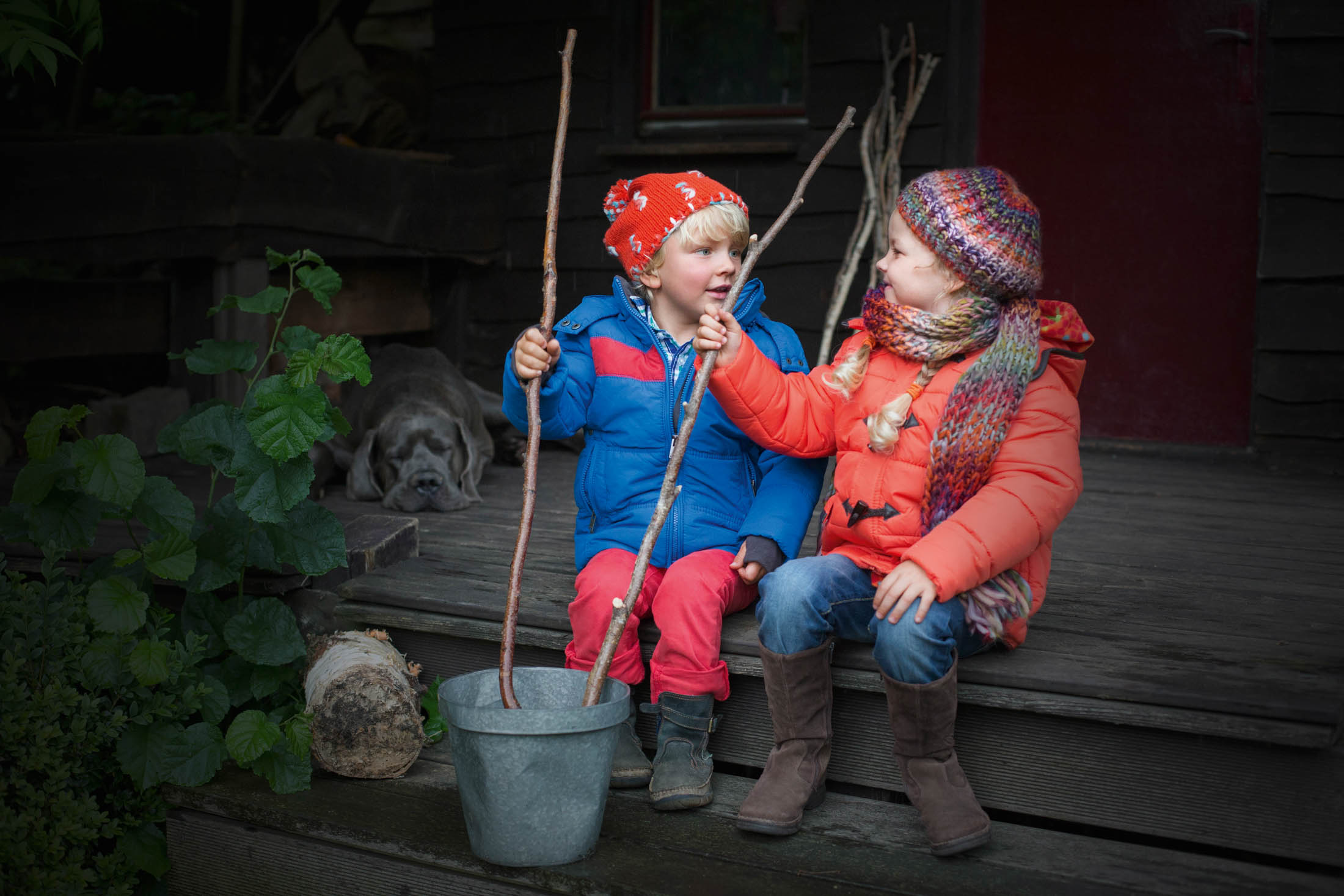 Zwei kleine Kinder in warmer Kleidung sitzen auf einer Holzveranda und halten spielerisch Stöcke in der Hand. Im Hintergrund liegt ein Hund bei einem Haus. Sie interagieren fröhlich miteinander. © Fotografie Tomas Rodriguez