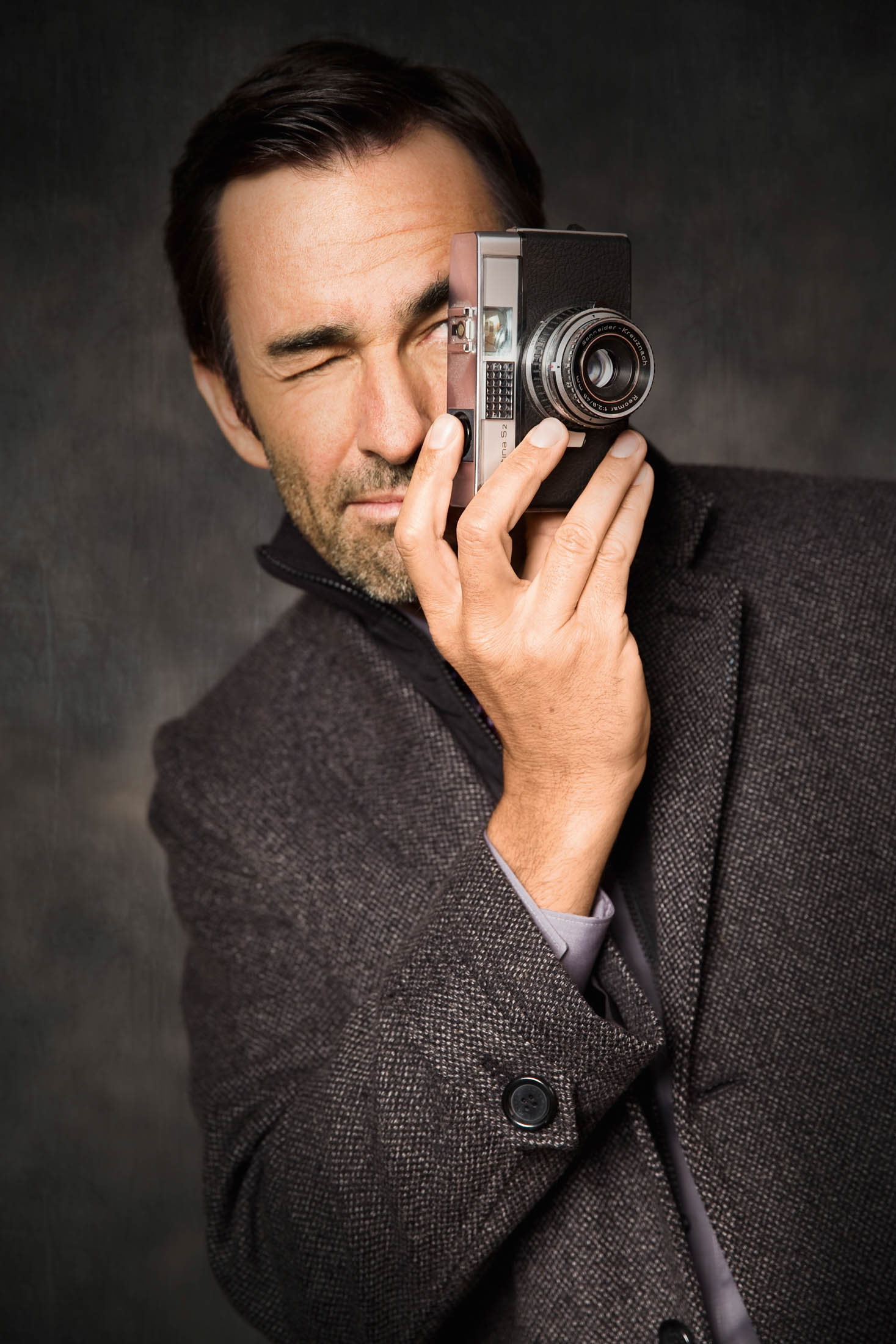 Ein Mann hält eine Vintage-Kamera vor sein Gesicht und fokussiert durch den Sucher. Er trägt einen dunklen Blazer und hat ein leichtes Grinsen. Er steht vor einem strukturierten grauen Hintergrund. © Fotografie Tomas Rodriguez
