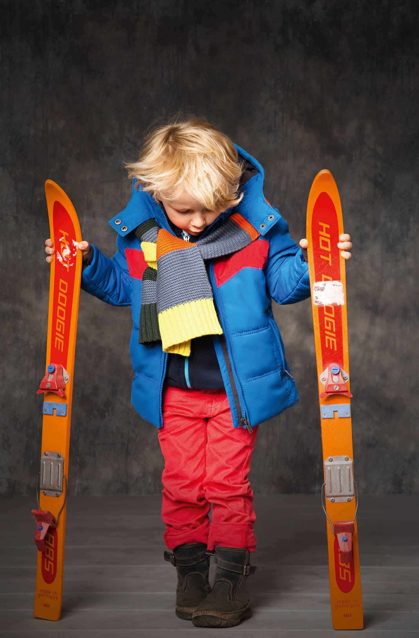Ein kleines Kind in einer bunten Winterjacke und roten Hosen hält ein Paar orangefarbene Skier in der Hand und blickt mit leichtem Stirnrunzeln auf sie herab, vor einem strukturierten grauen Hintergrund. © Fotografie Tomas Rodriguez