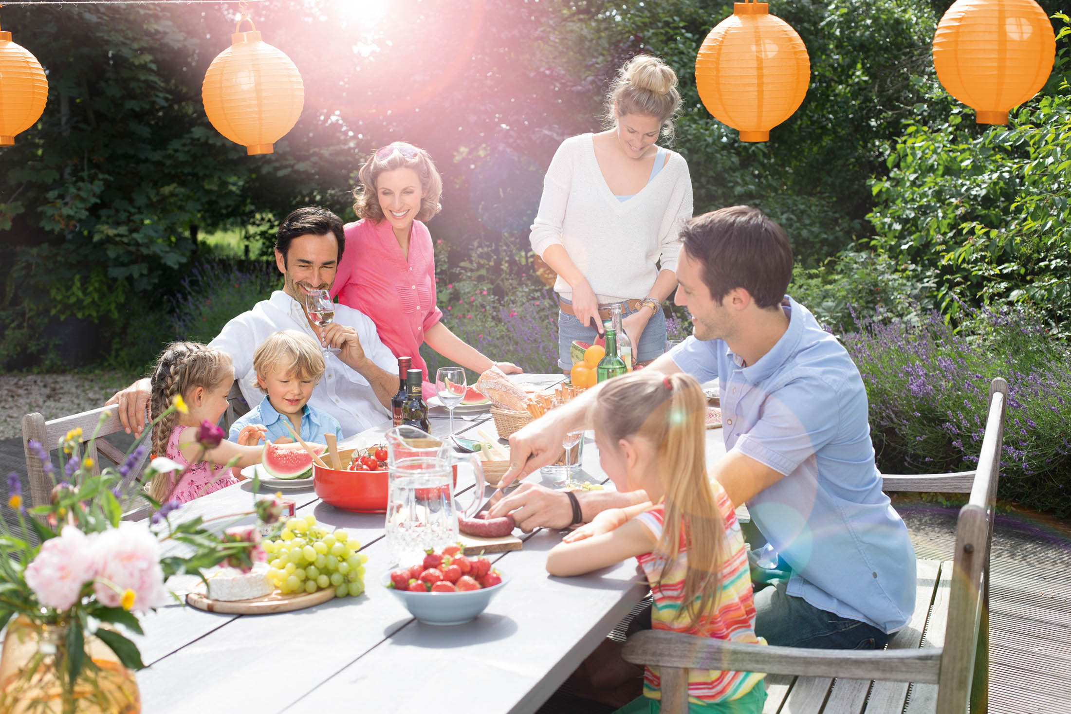Eine Mehrgenerationenfamilie genießt ein Essen im Freien in einem mit orangefarbenen Laternen geschmückten Garten. Die Kinder essen und die Erwachsenen lächeln, umgeben von Natur und sonniger Atmosphäre. © Fotografie Tomas Rodriguez