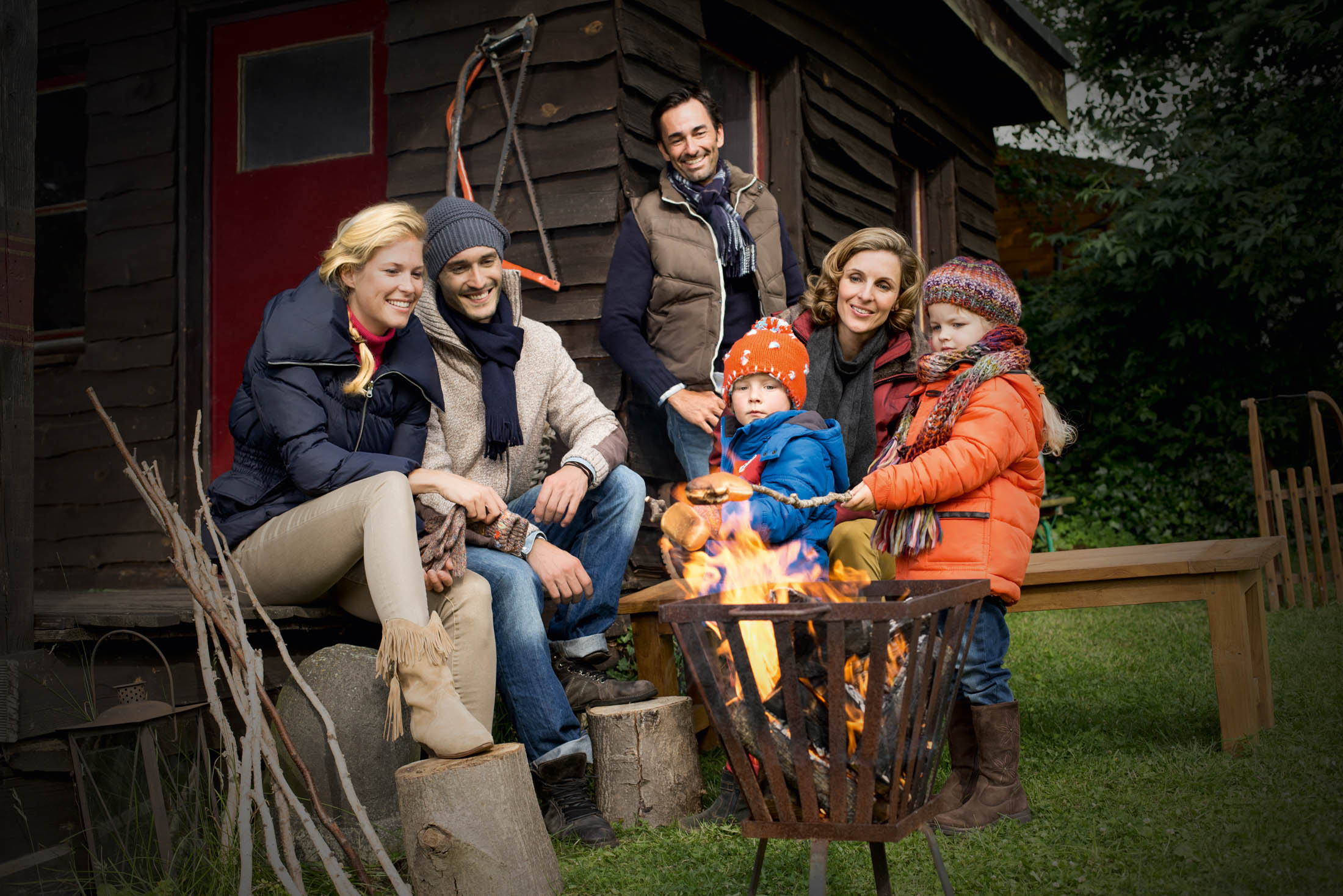 Zwei Familien genießen die Zeit an einer Feuerstelle vor einer Hütte. Die Erwachsenen lächeln, die Kinder sind warm angezogen. Natürliche Umgebung mit grünem Gras und Bäumen im Hintergrund. © Fotografie Tomas Rodriguez