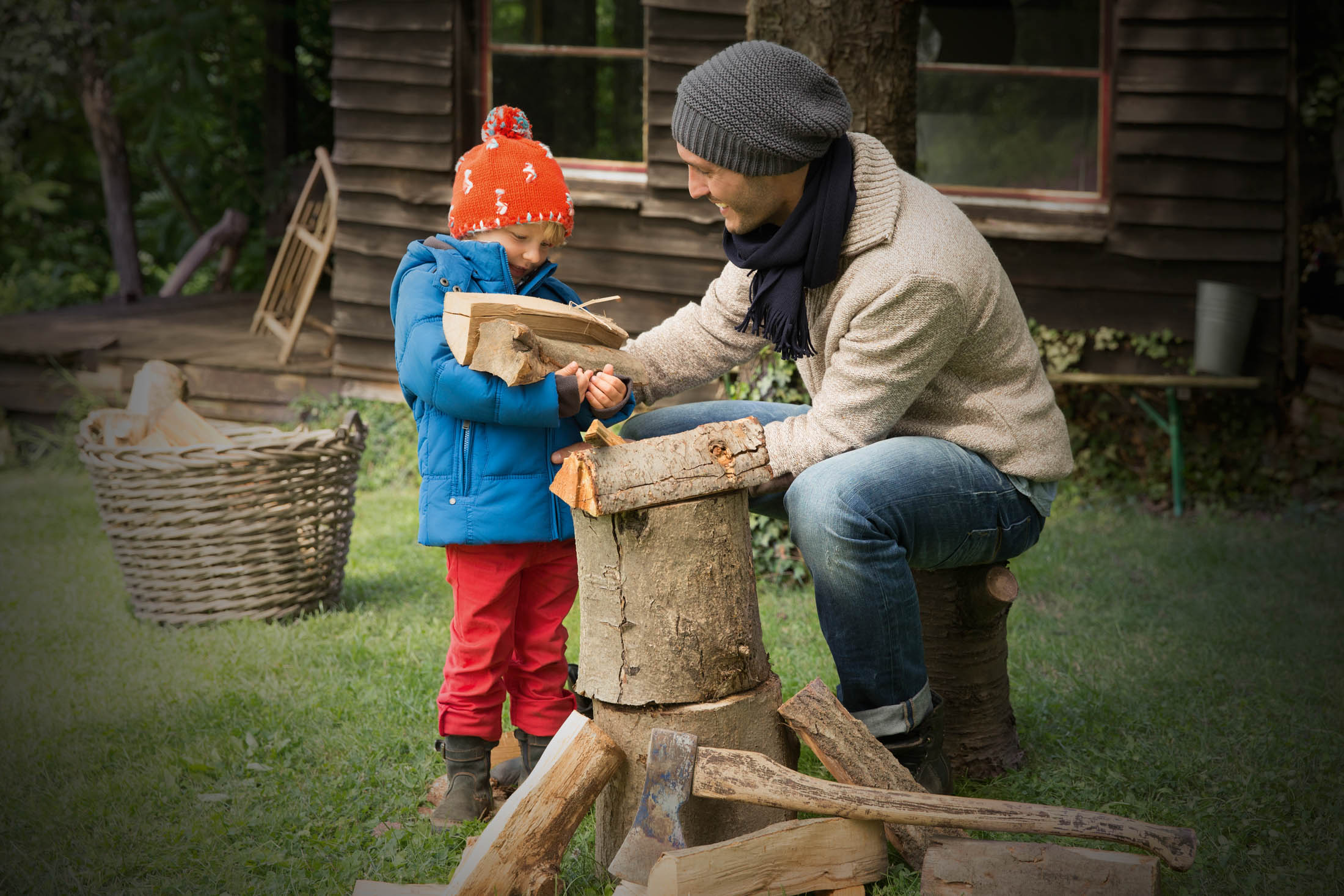 Ein Mann mit Mütze und Schal bringt einem kleinen Kind mit einer leuchtend orangefarbenen Mütze bei, wie man mit einer kleinen Axt Holz hackt. In der Nähe einer rustikalen Holzhütte und eines Korbes mit Holzstämmen. © Fotografie Tomas Rodriguez