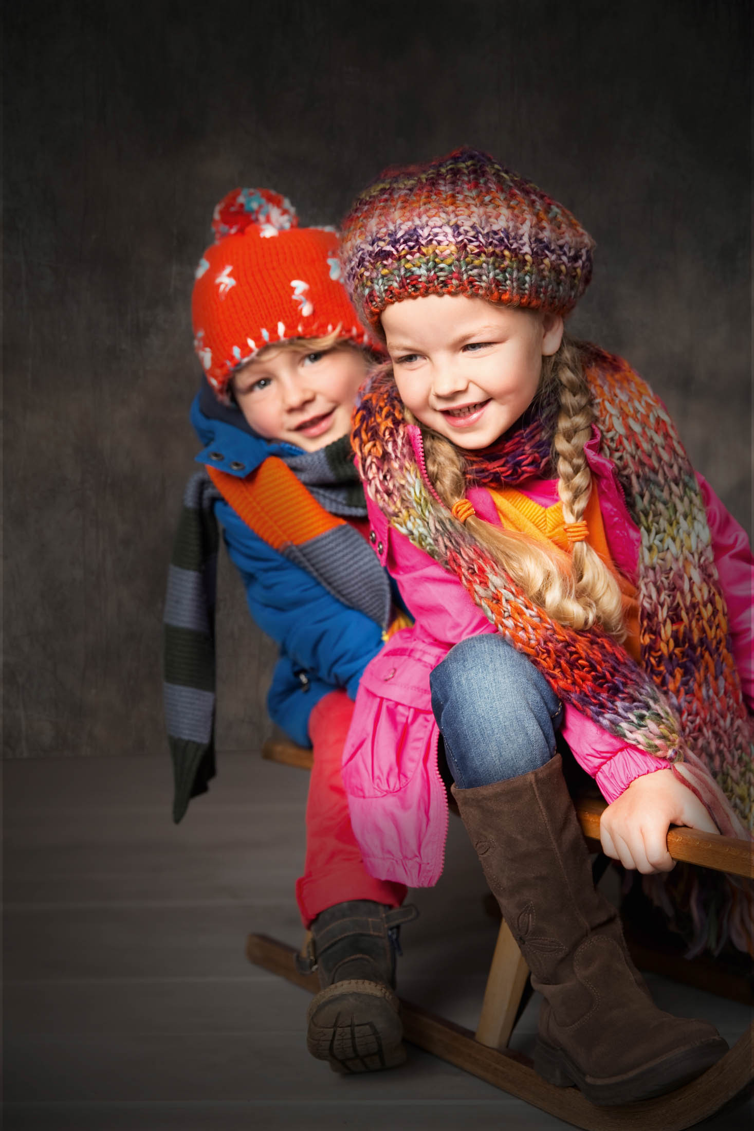 Zwei Kinder in Winterkleidung, darunter bunte Mützen und Schals, sitzen lächelnd zusammen auf einem Holzschlitten vor einem grauen Hintergrund. © Fotografie Tomas Rodriguez