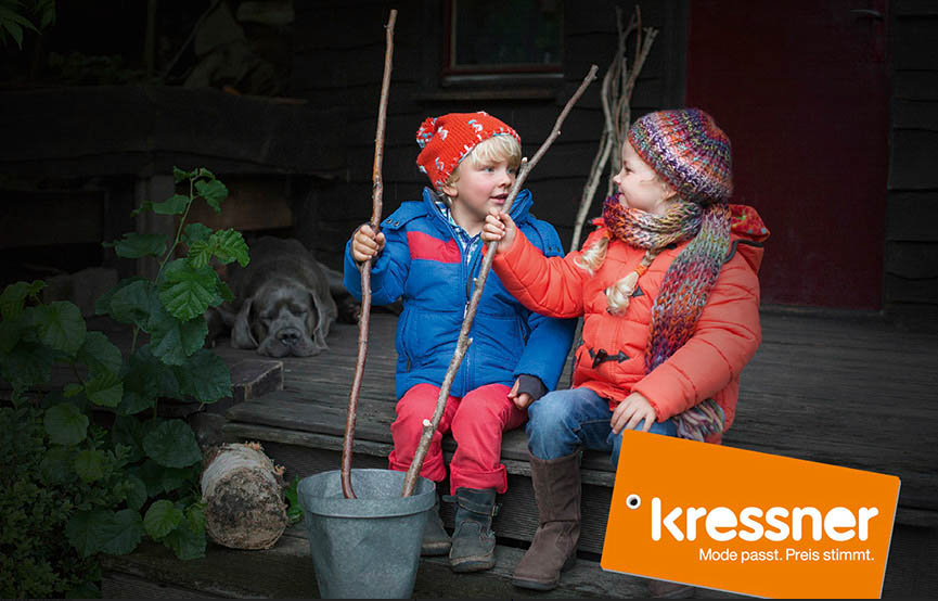 Zwei kleine Kinder in Winterkleidung spielen mit Stöcken auf einer Veranda, im Hintergrund schläft ein Hund und in der Ecke steht ein Logo mit der Aufschrift „Kressner“. © Fotografie Tomas Rodriguez