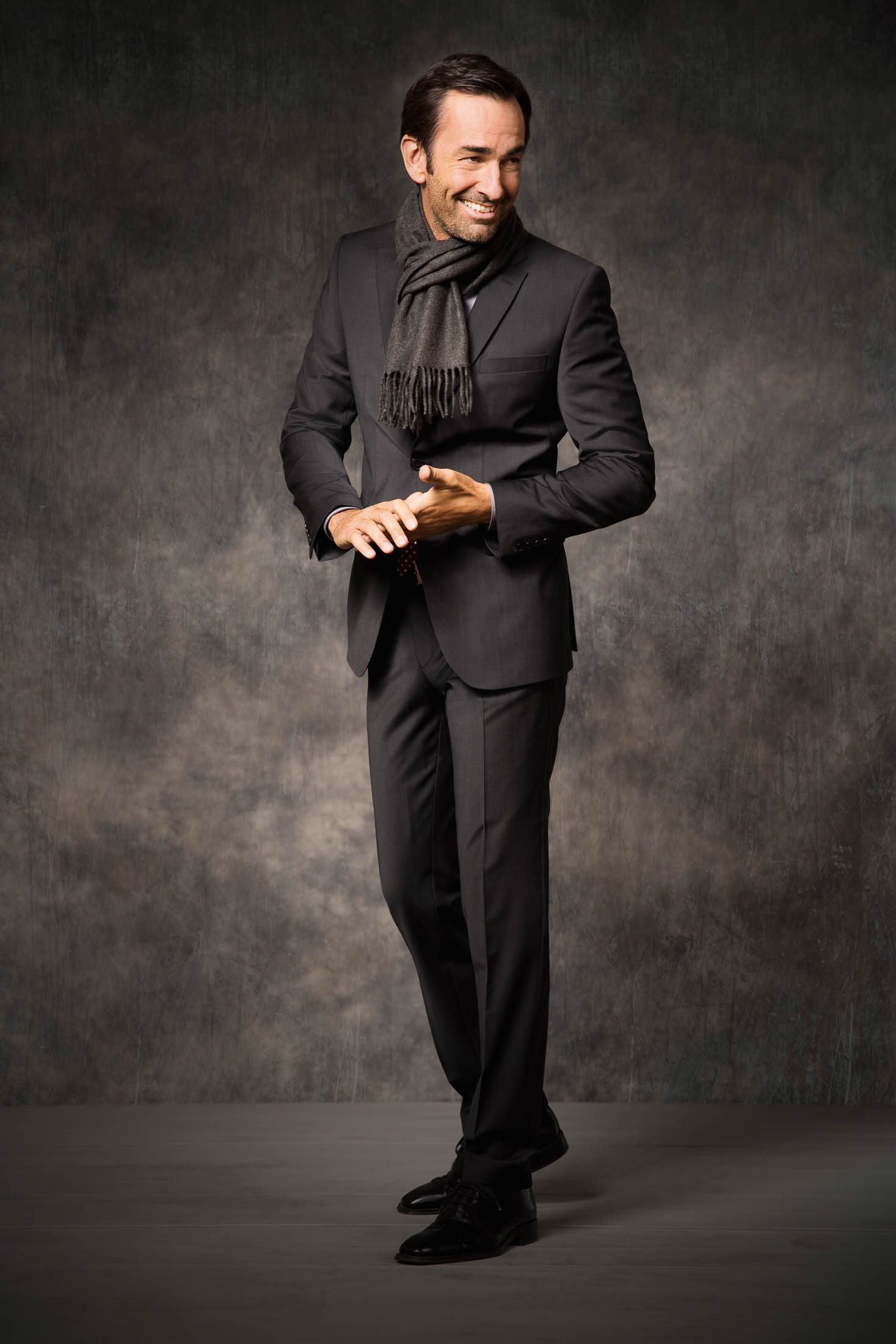 Eleganter Mann in maßgeschneidertem Anzug und Schal, lächelnd und die Hände faltend, steht vor einem strukturierten grauen Hintergrund. © Fotografie Tomas Rodriguez